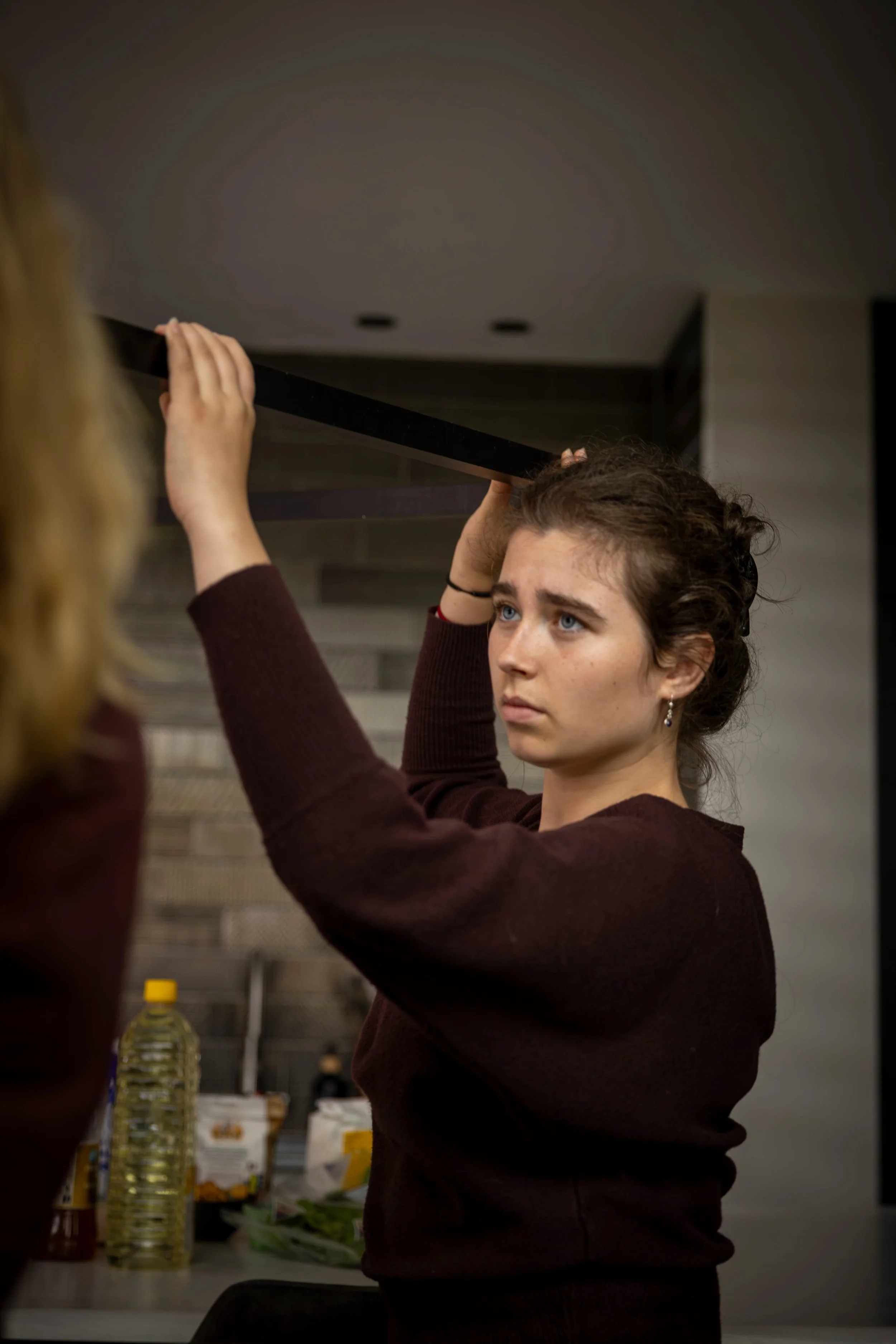 Young woman with brown hair and blue eyes, wearing a maroon sweater, lifting a black shelf or large object in a kitchen or dining area.