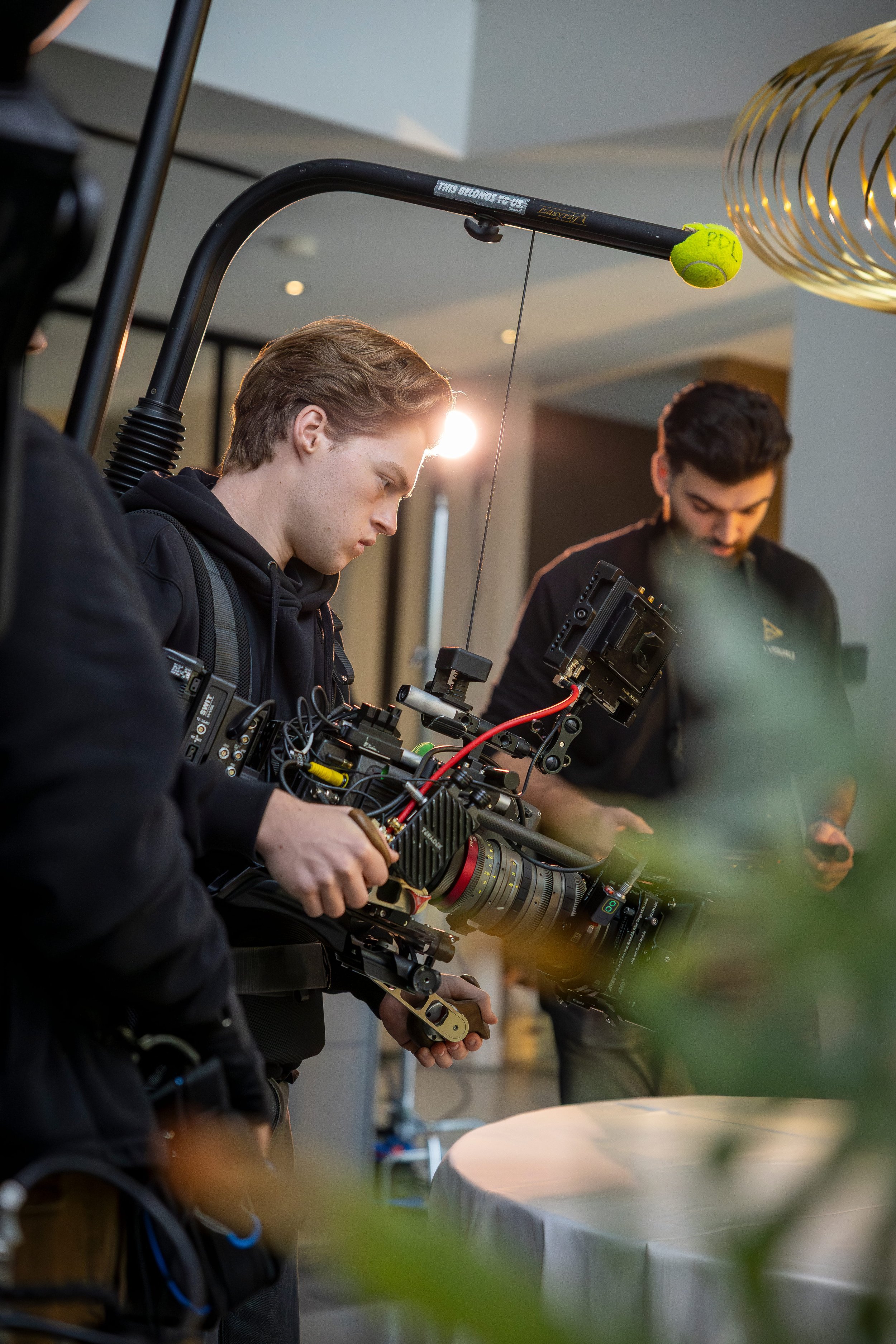 Filmmaker operating a professional camera with a large lens and monitor in an indoor setting, with a crew member using a smartphone nearby.