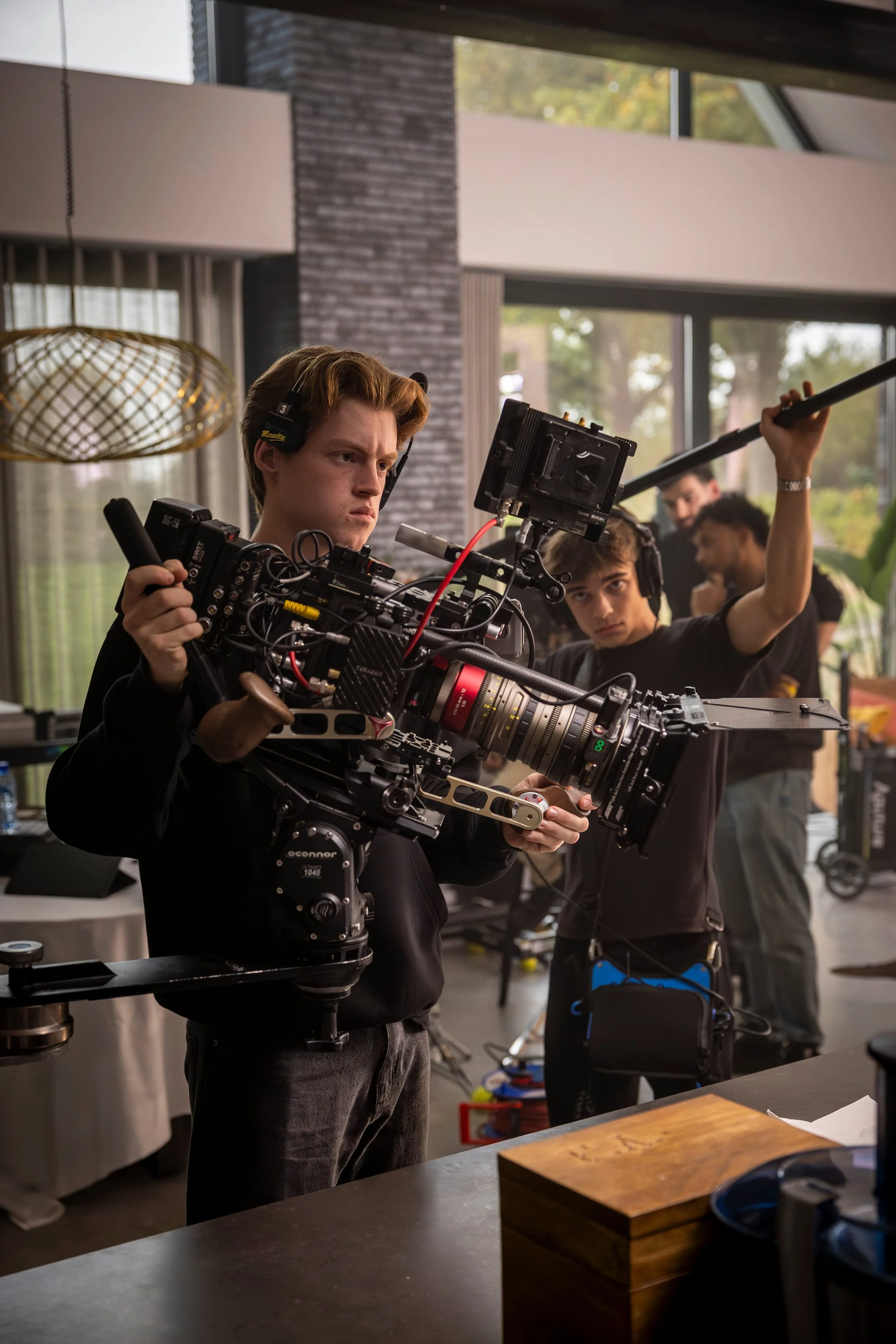 A young man operating a professional camera on a film set, with other crew members in the background, indoors with large windows and modern decor.