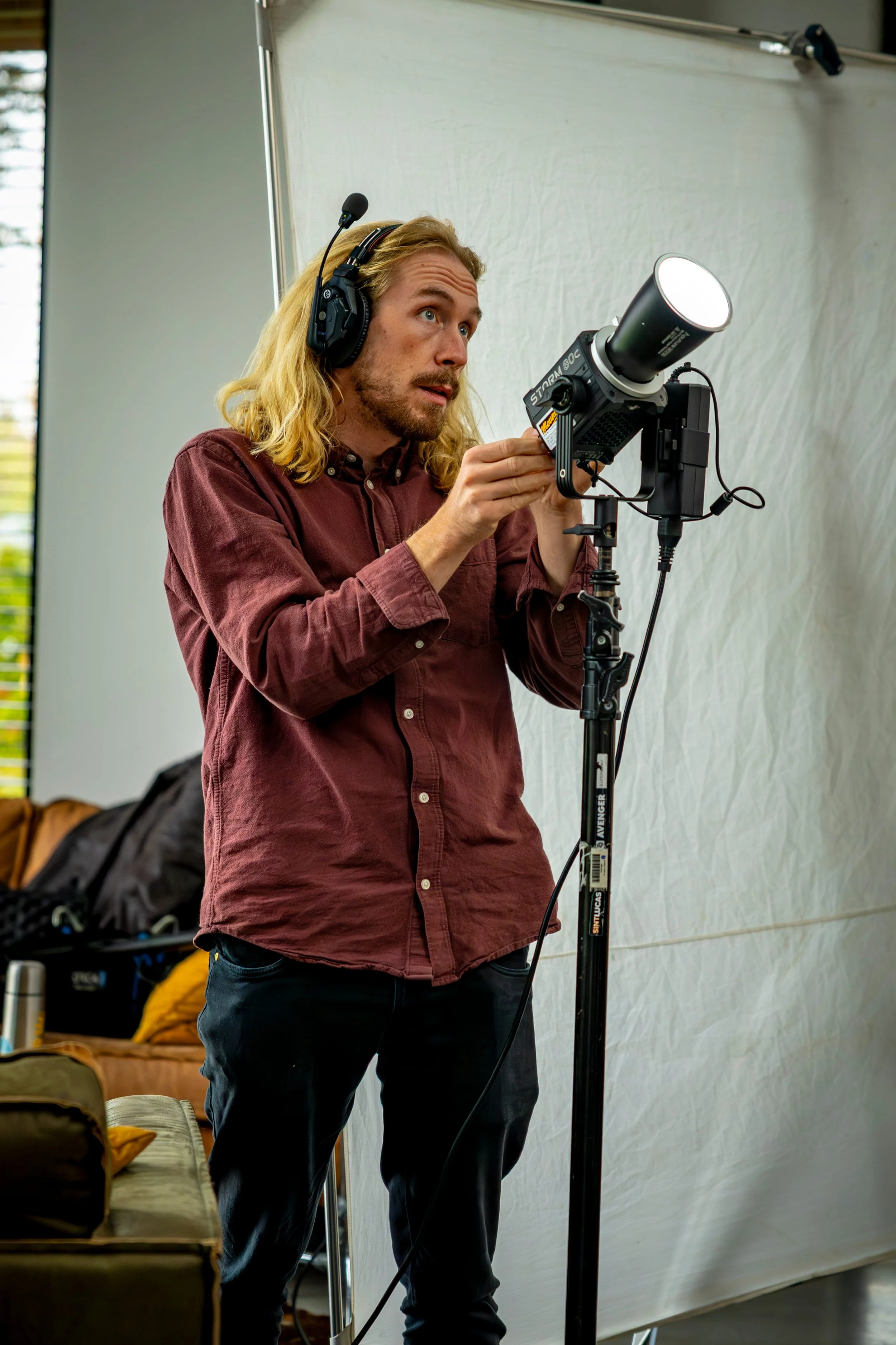 A man with long blonde hair, wearing a maroon shirt, adjusting a camera or telescope with headphones on, in front of a white backdrop.
