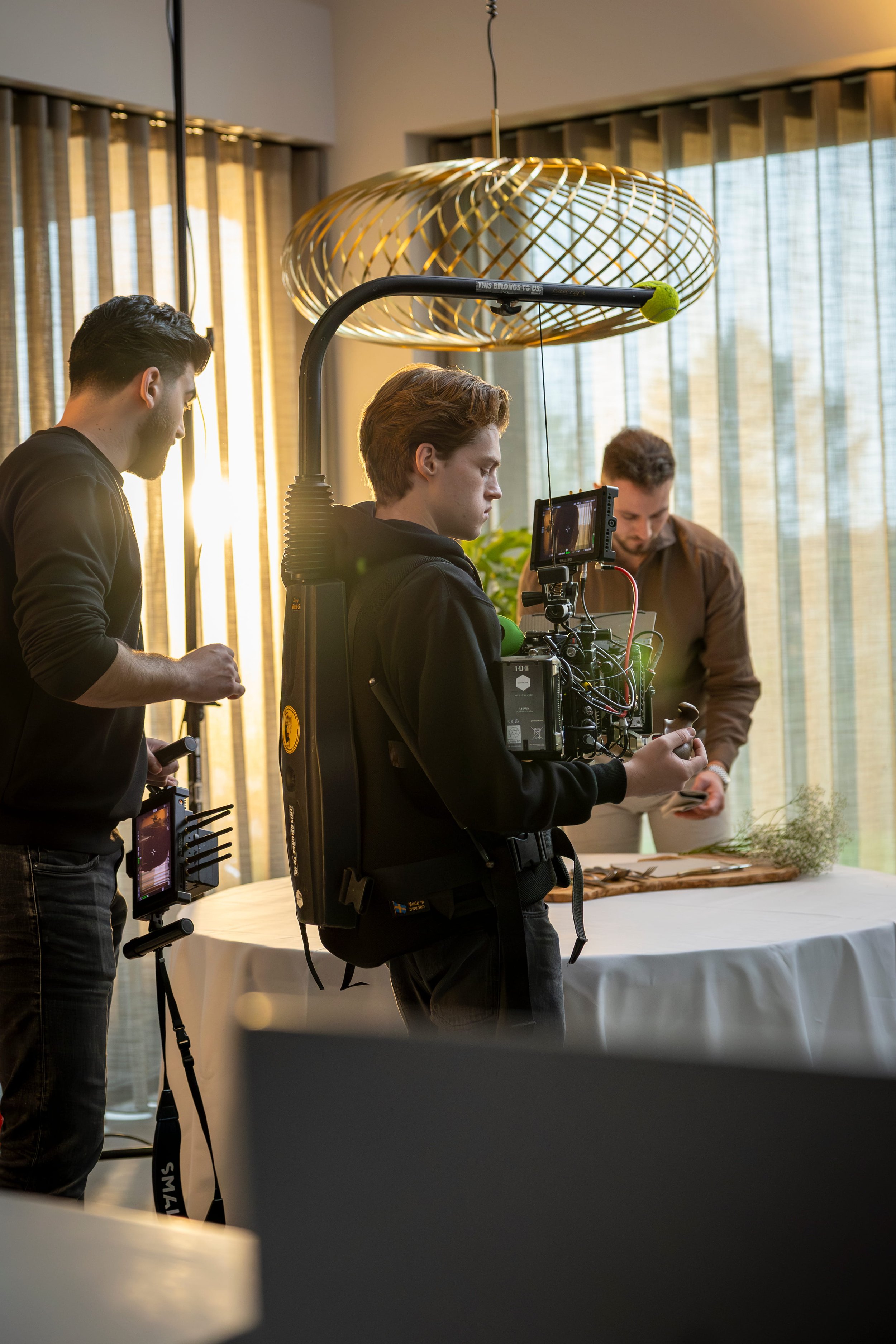 Film crew filming a scene indoors with a camera, monitor, and lighting equipment pointed at a table with utensils and greenery, near large windows with vertical blinds.