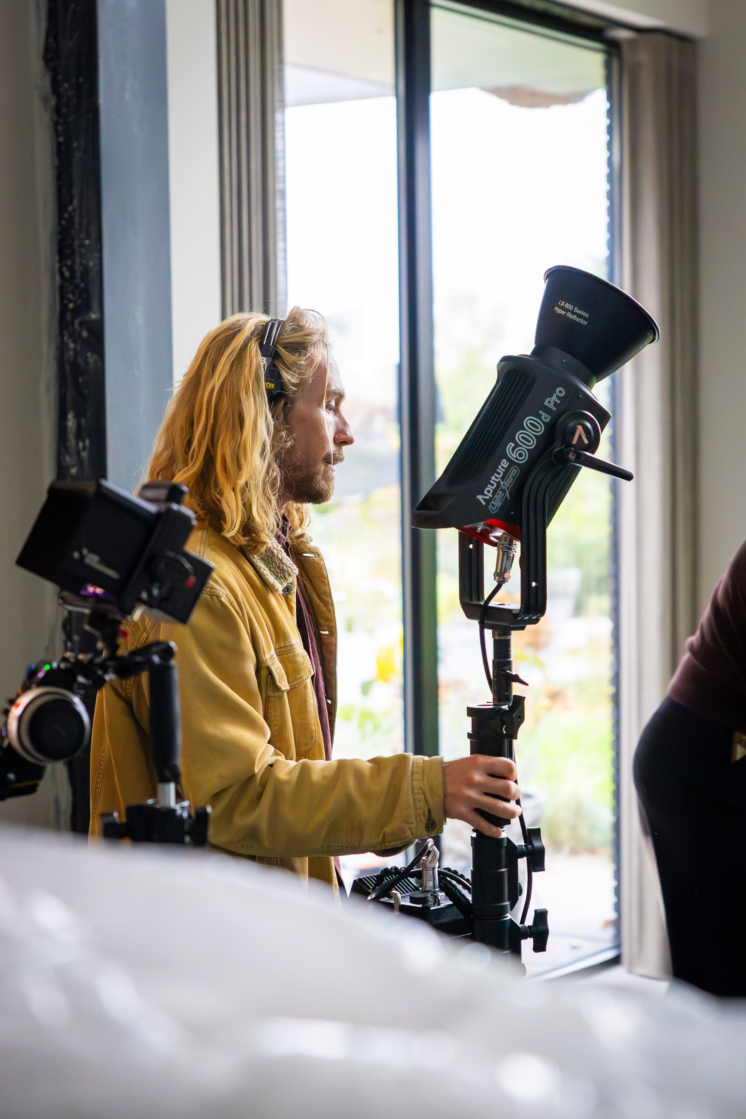 A man with long blond hair and a beard operating a large camera on a tripod indoors, with a window in the background.