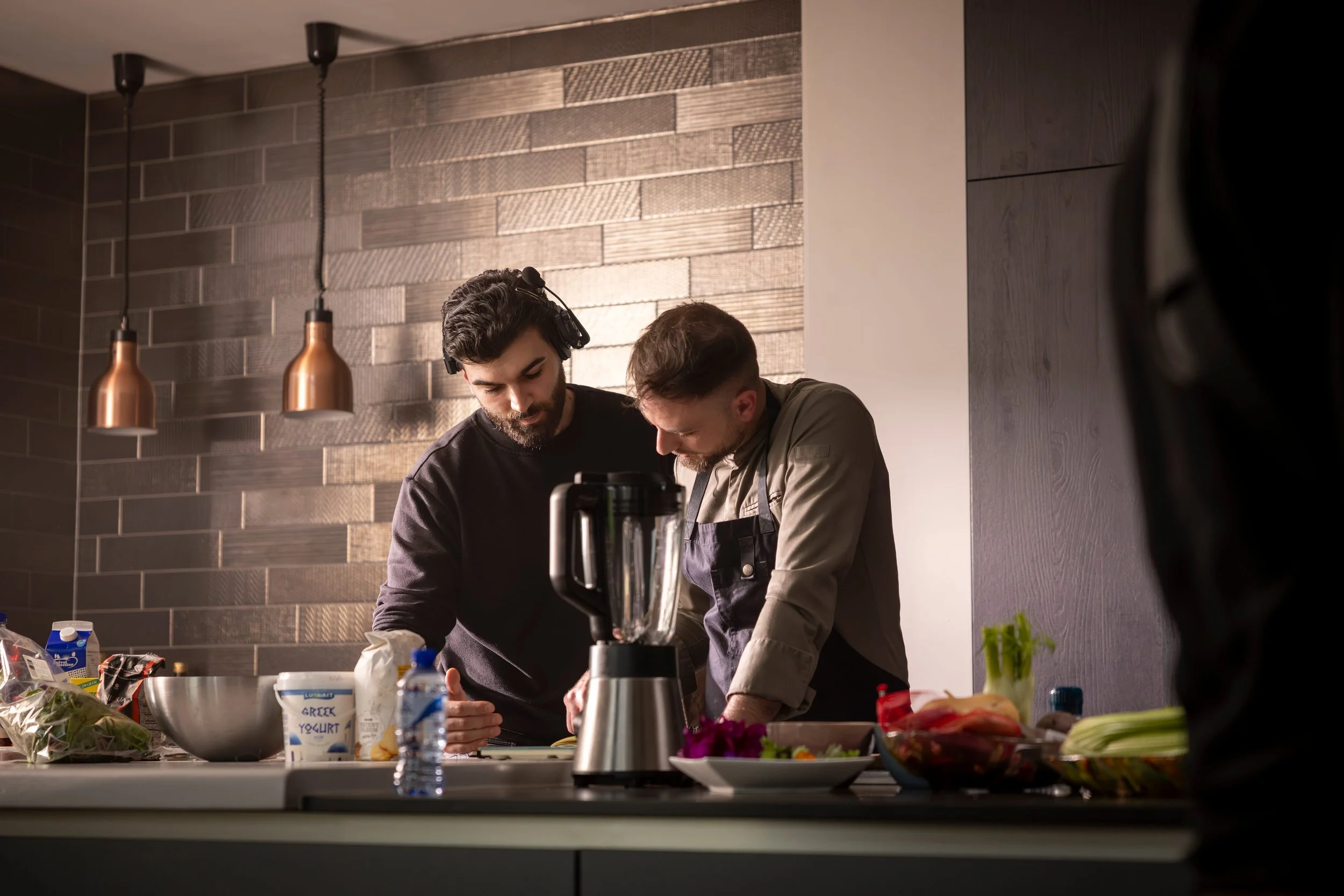 Two men cooking in a modern kitchen with wooden accent wall, one wearing headphones, the other in an apron, surrounded by ingredients and a blender.