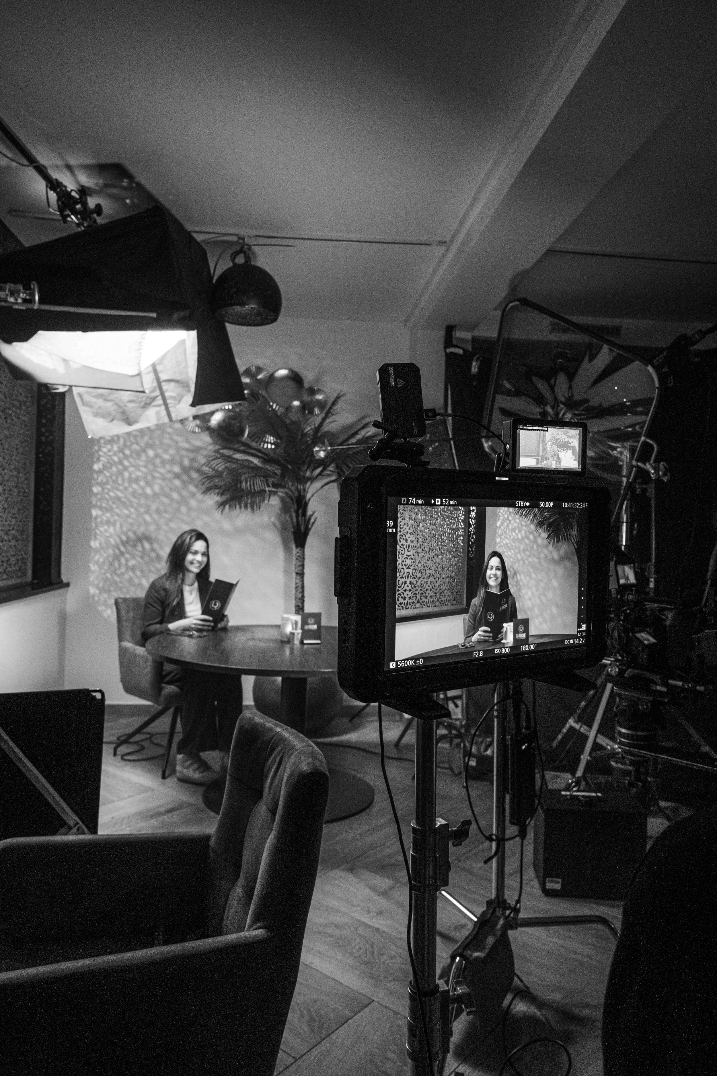 A woman sitting at a round table inside a studio, looking at the camera with a smile, holding a document. The scene is being filmed with professional camera equipment, and there is a large plant and textured wall in the background.