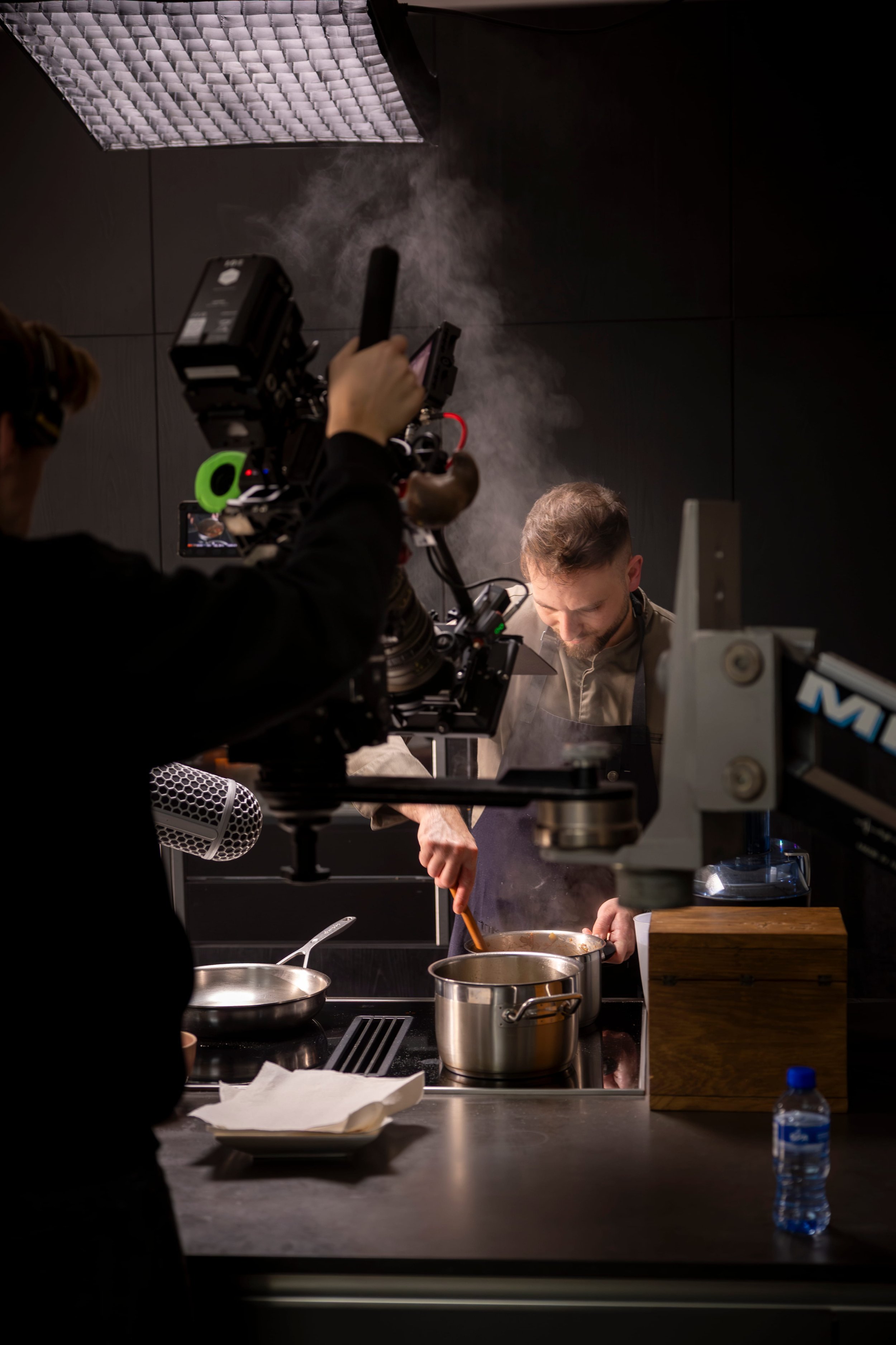 A chef cooking in a professional kitchen with a video camera operator filming his actions, steam rising from pots on the stove, and kitchen utensils on the counter.