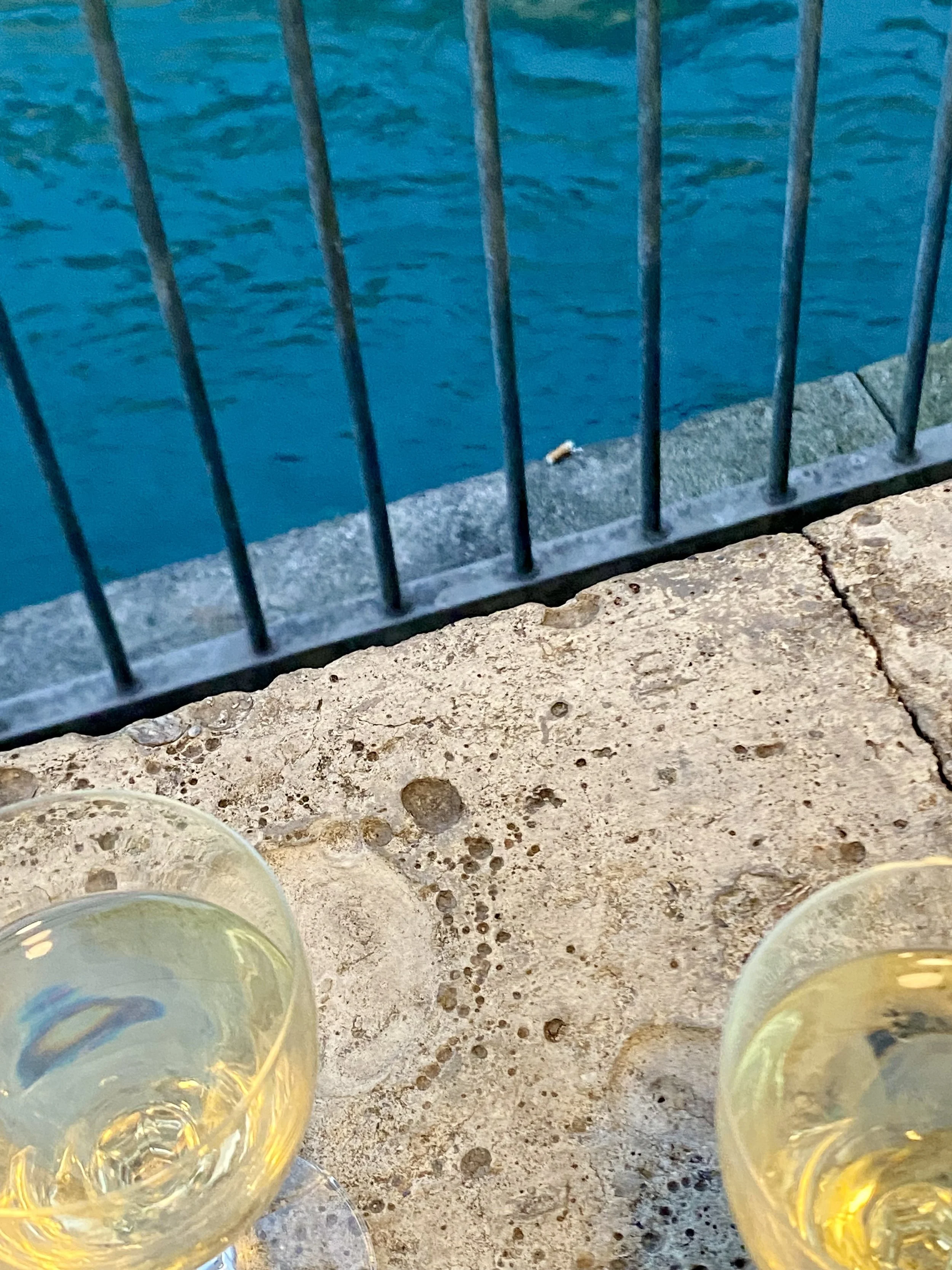 Zwei Weingläser auf einer Steinbrüstung vor blauem Wasser und einem Geländer.