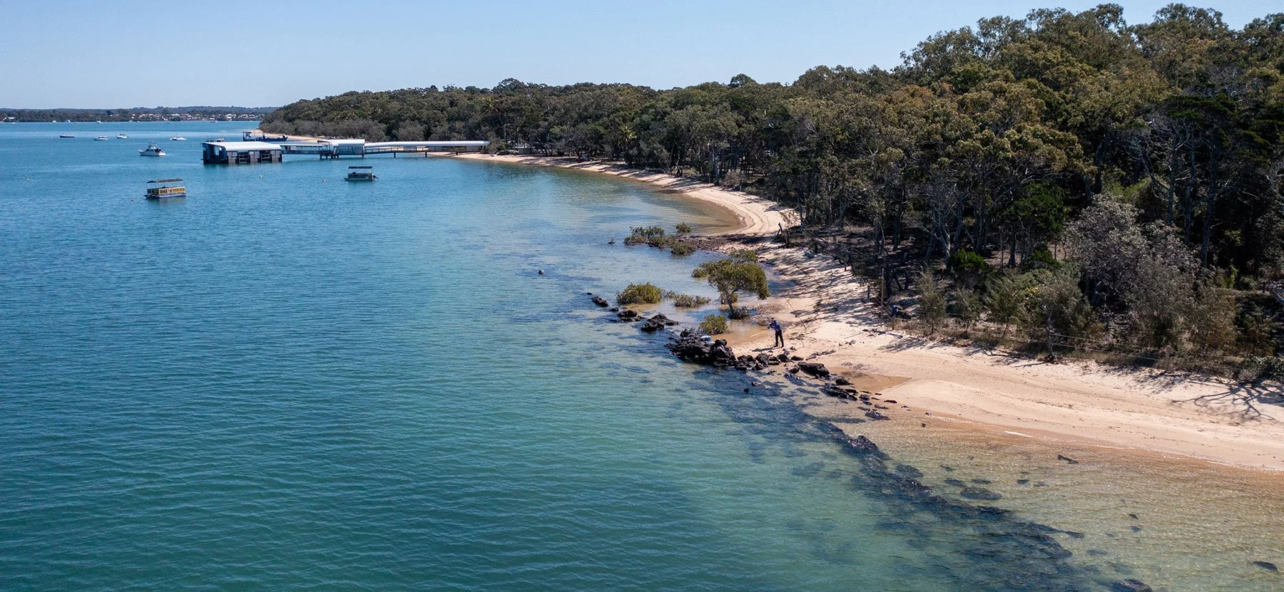 A peaceful coastal scene with a sandy beach, trees along the shoreline, and boats on calm water near a pier.