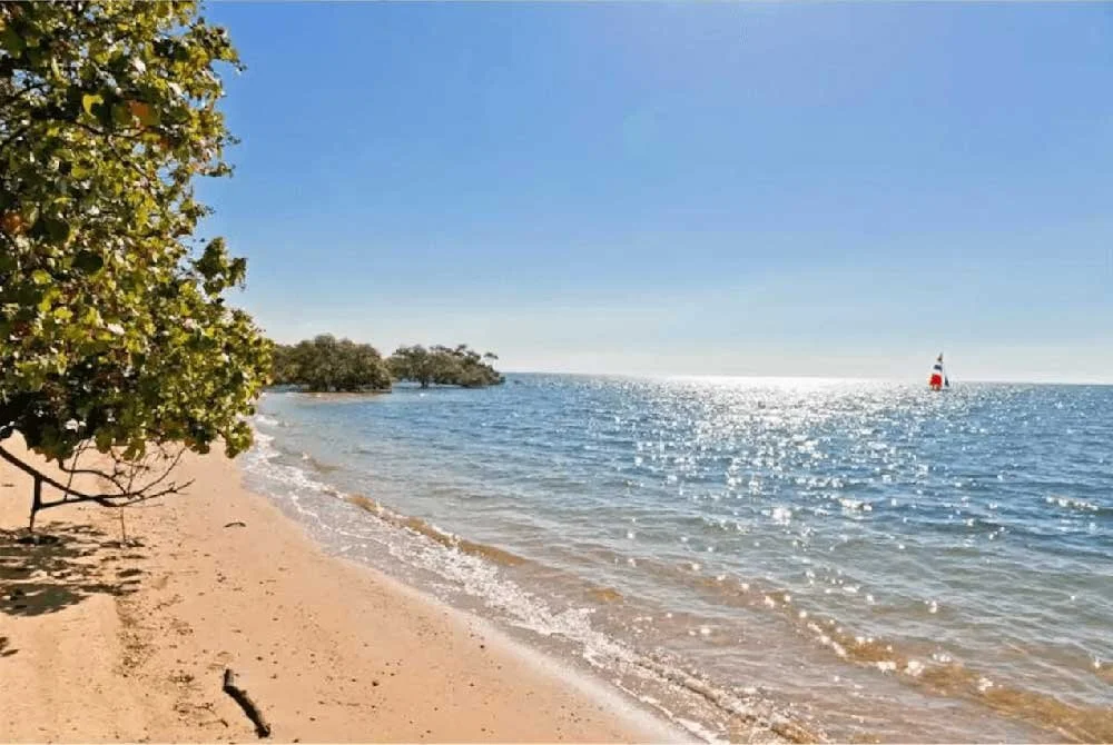 Sandy beach, glistening bay with a yacht on the horizon with red, blue and white sails.