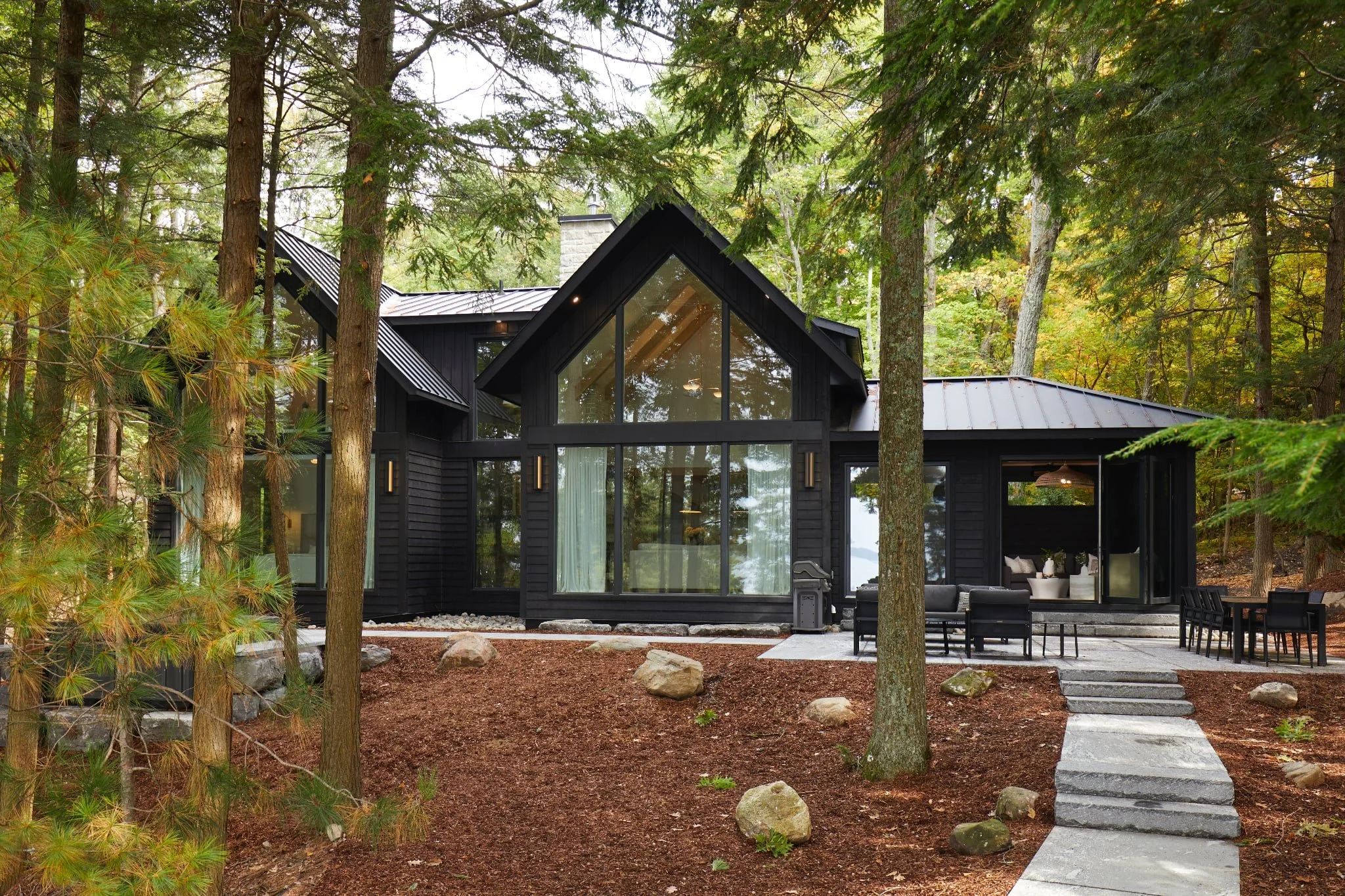 Black modern house with large glass front, surrounded by trees in a forest setting, with stone steps leading to a patio area.