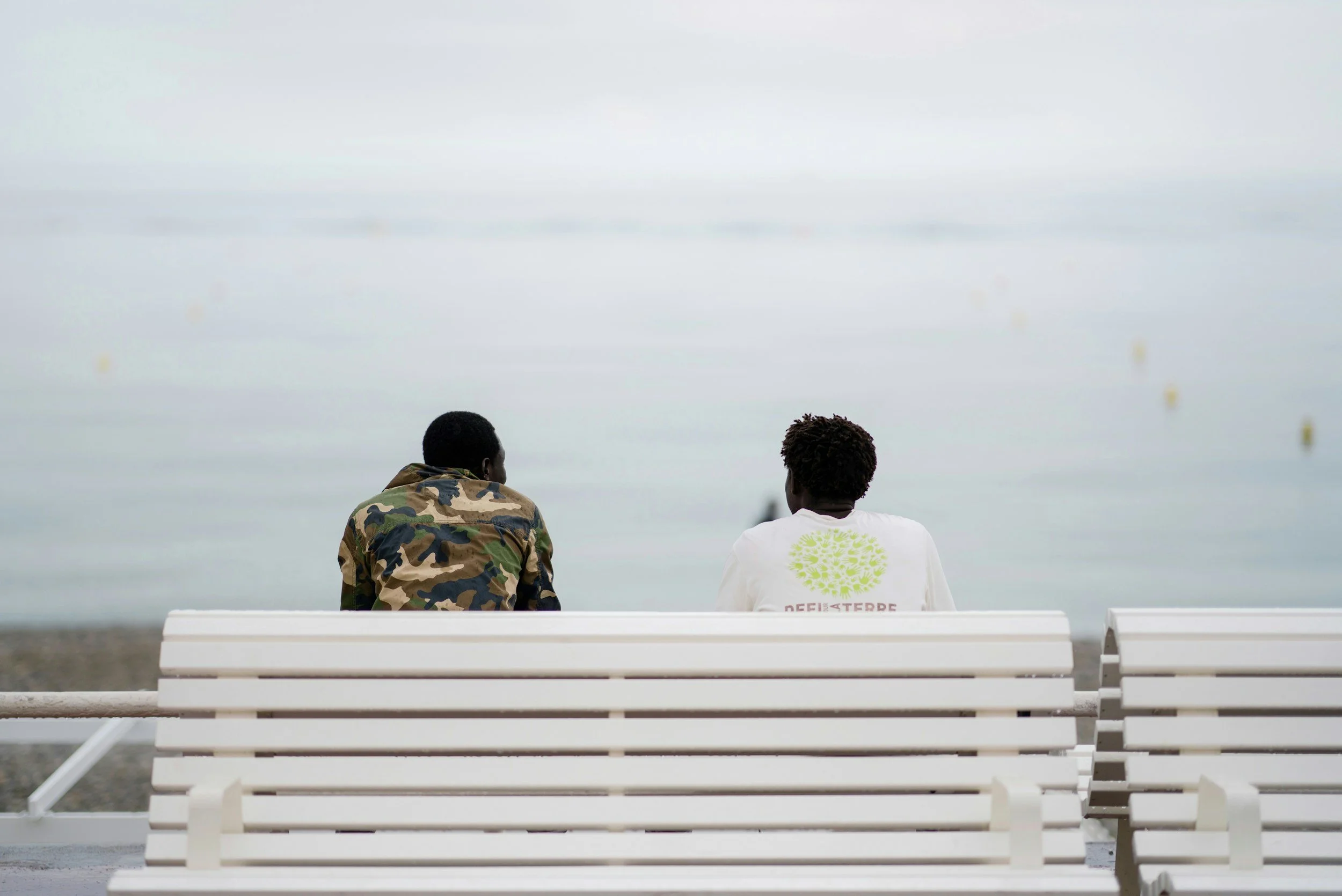 Two men sitting on a white bench by the water, facing away from the camera, with a cloudy sky in the background.