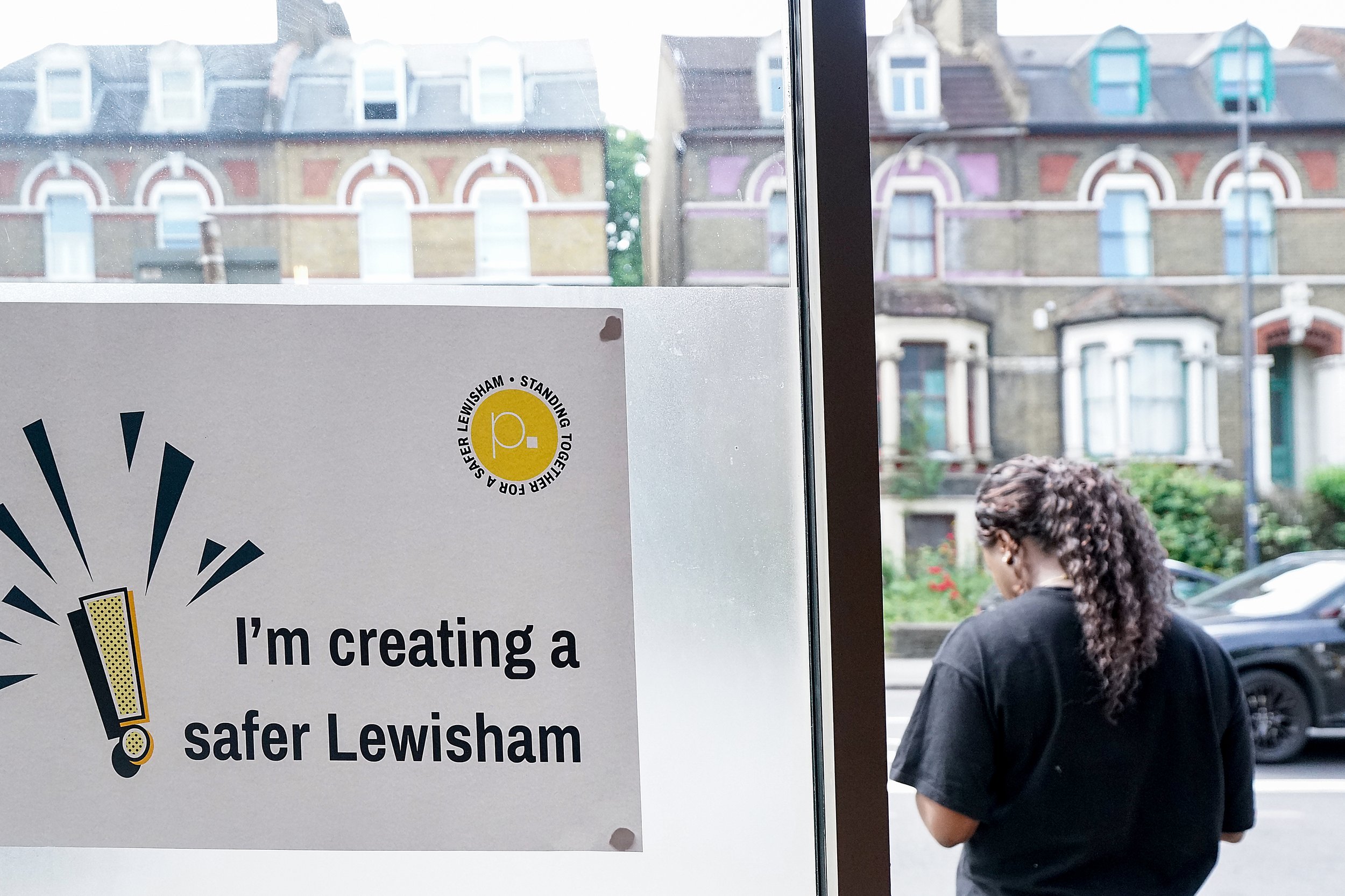A woman with curly hair standing outside on a city street, looking to the side. There is a poster on the glass door that says 'I'm creating a safer Lewisham' with a yellow circle logo.