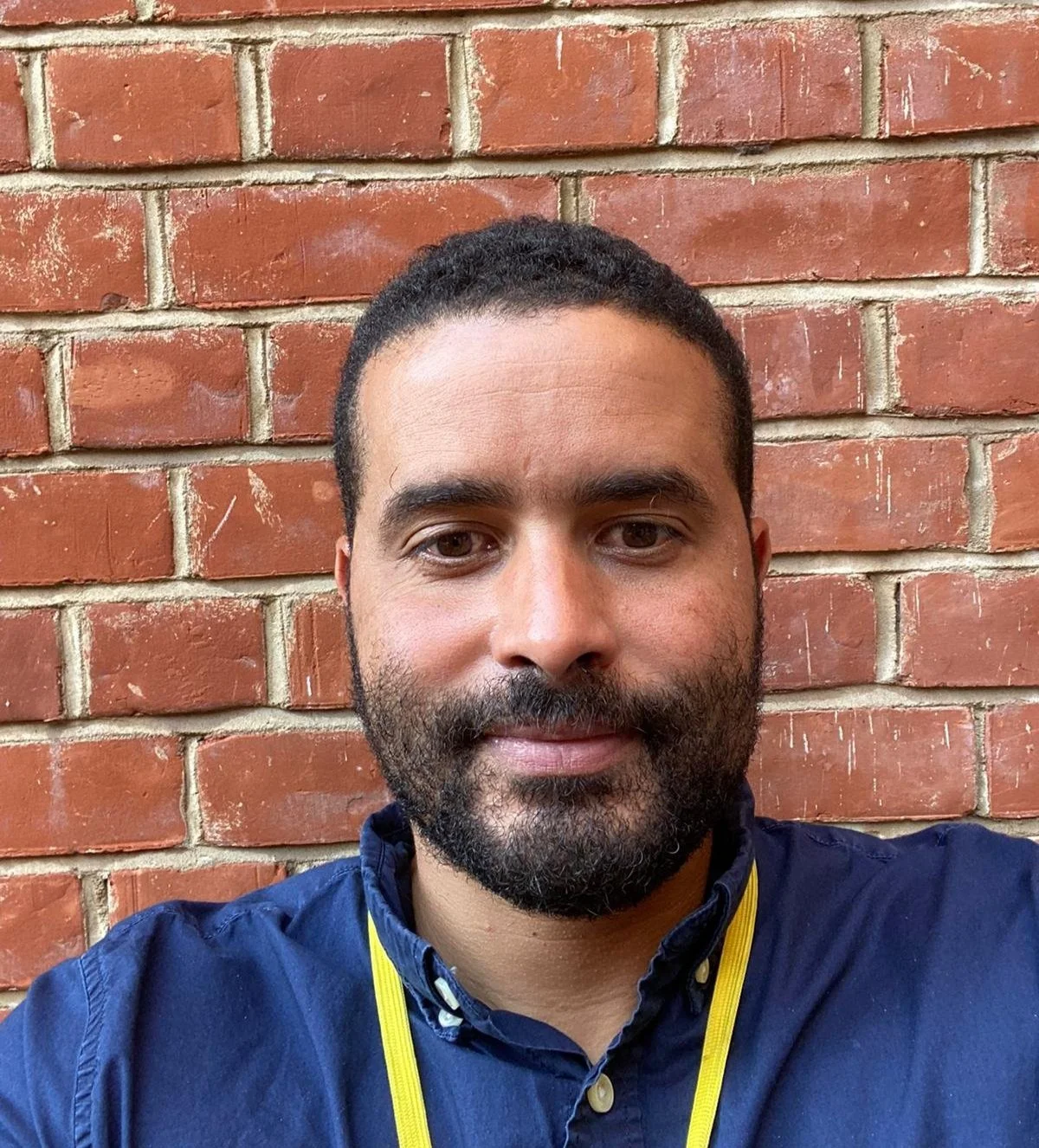 Portrait of a person with a beard wearing a blue shirt and yellow lanyard, standing against a red brick wall.