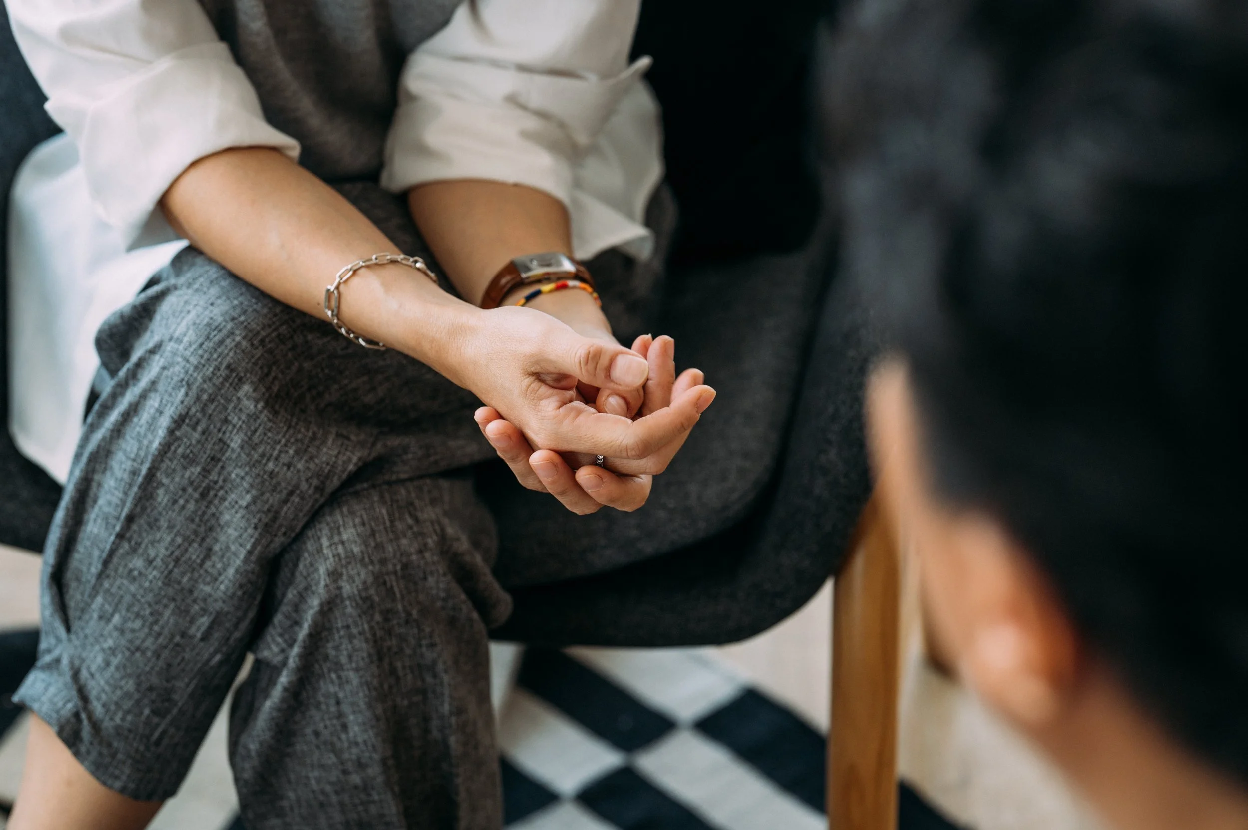 A person wearing a long-sleeve white shirt and gray checked pants sitting on a dark sofa with their hands clasped together on their lap.