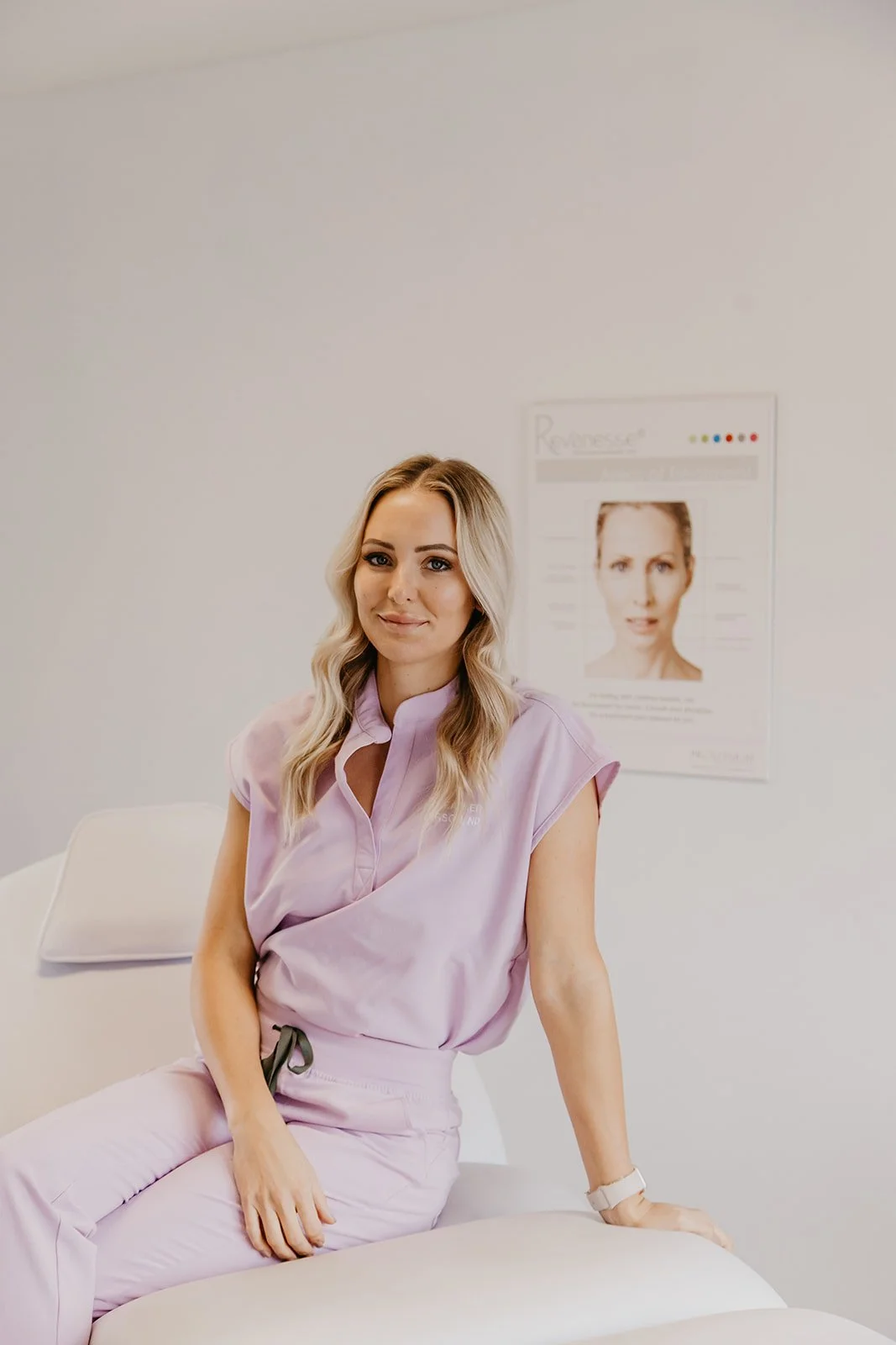 A woman in a lilac uniform sits on a medical examination chair in a clinic room with a poster on the wall.