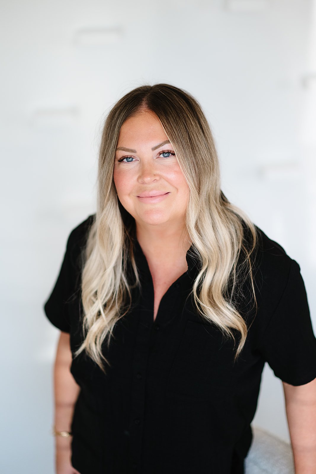 Person with long blonde hair wearing a black top, sitting in front of a light-colored wooden wall.