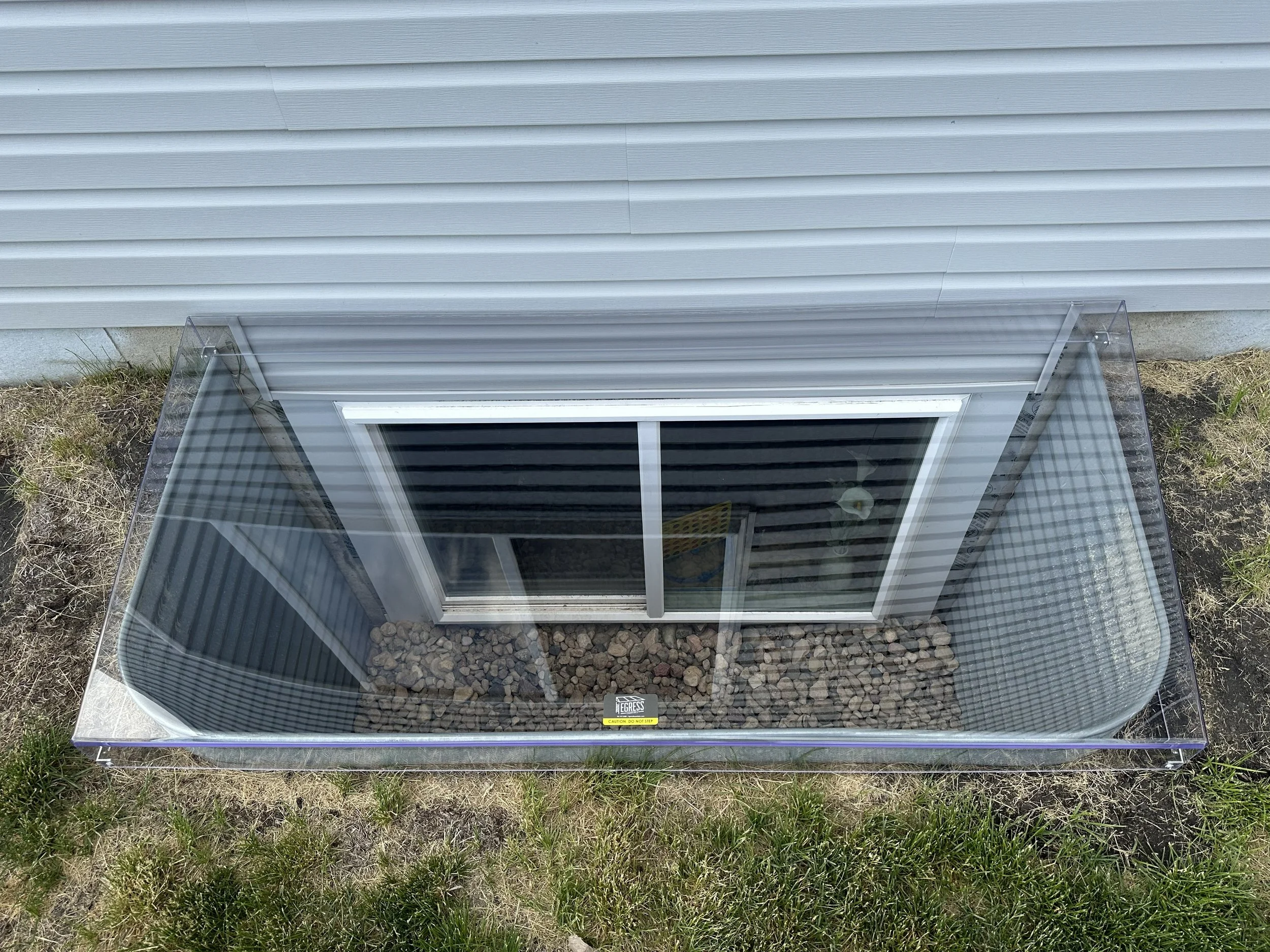 A square glass enclosure with rocks inside, positioned on grass outside a building with beige siding. A window is visible in the enclosure, and a caution sign is attached to the glass at the bottom.