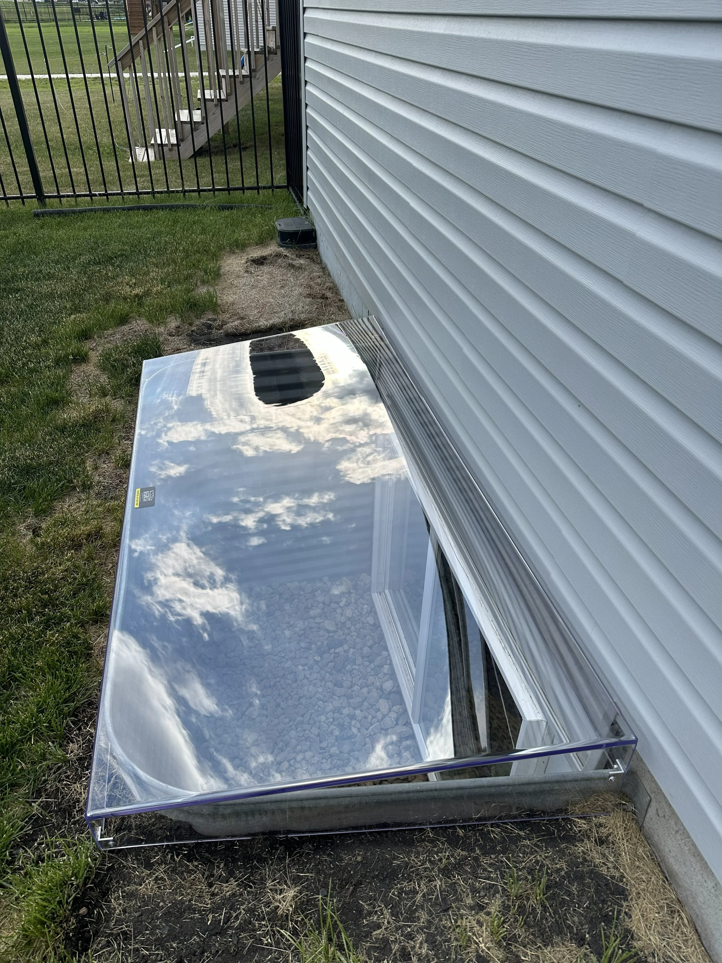 A small glass terrarium with a reflective top, placed on the ground next to a white house wall, with a green grassy area and a black metal fence in the background. The reflection on the glass shows a cloudy sky.