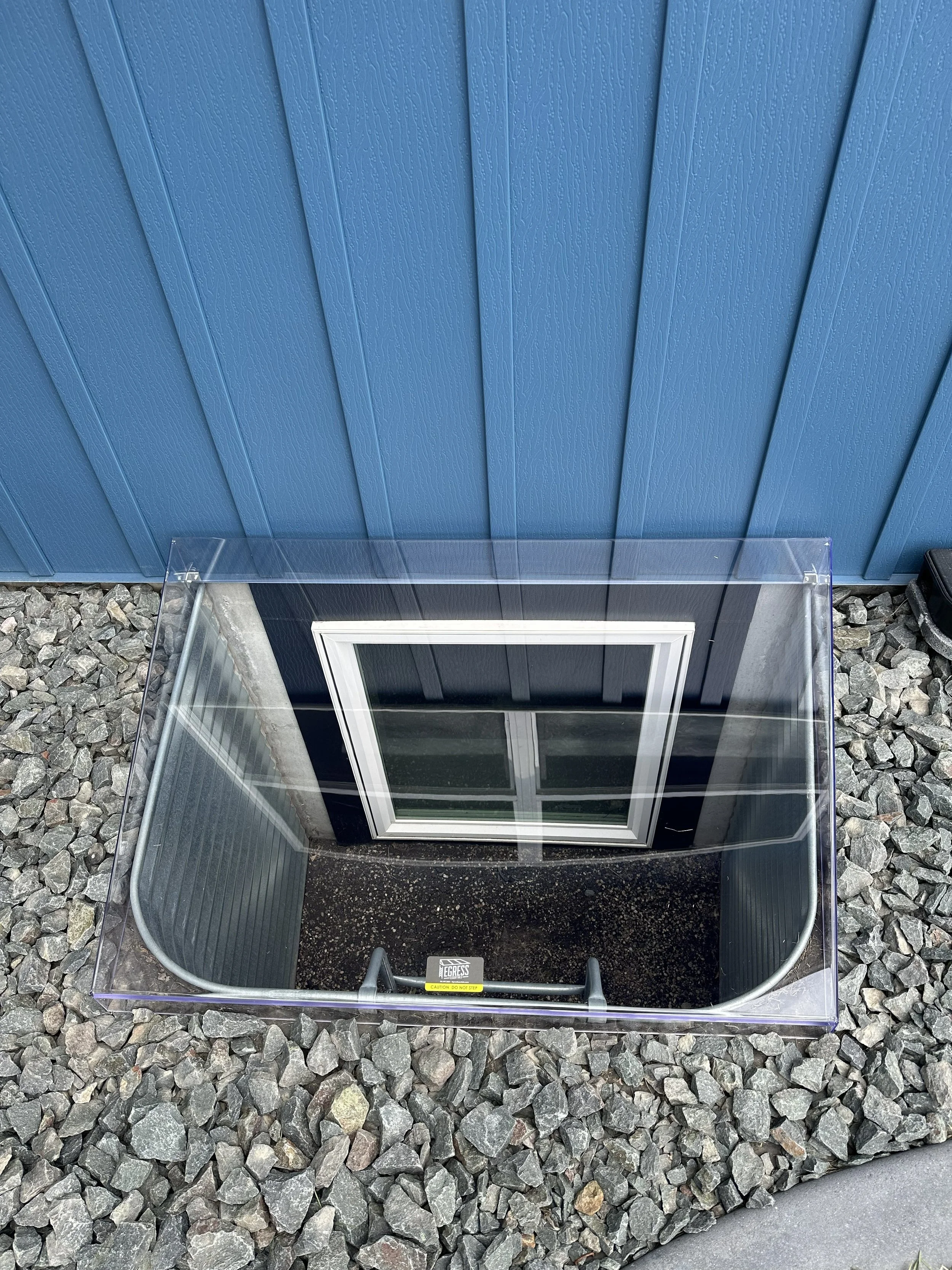 An outdoor scene showing a vent or exhaust fan on a blue building wall, surrounded by gravel, with a transparent enclosure and a caution sign on the enclosure.