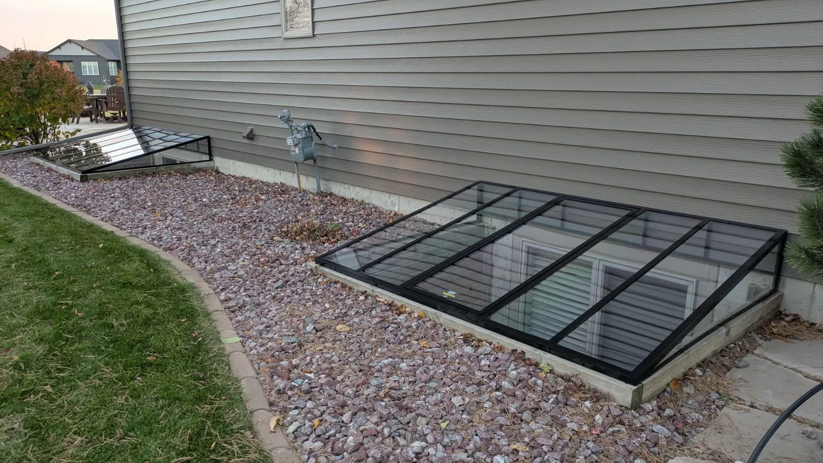 Raised garden beds with glass covers on a gravel bed next to a house with gray siding.
