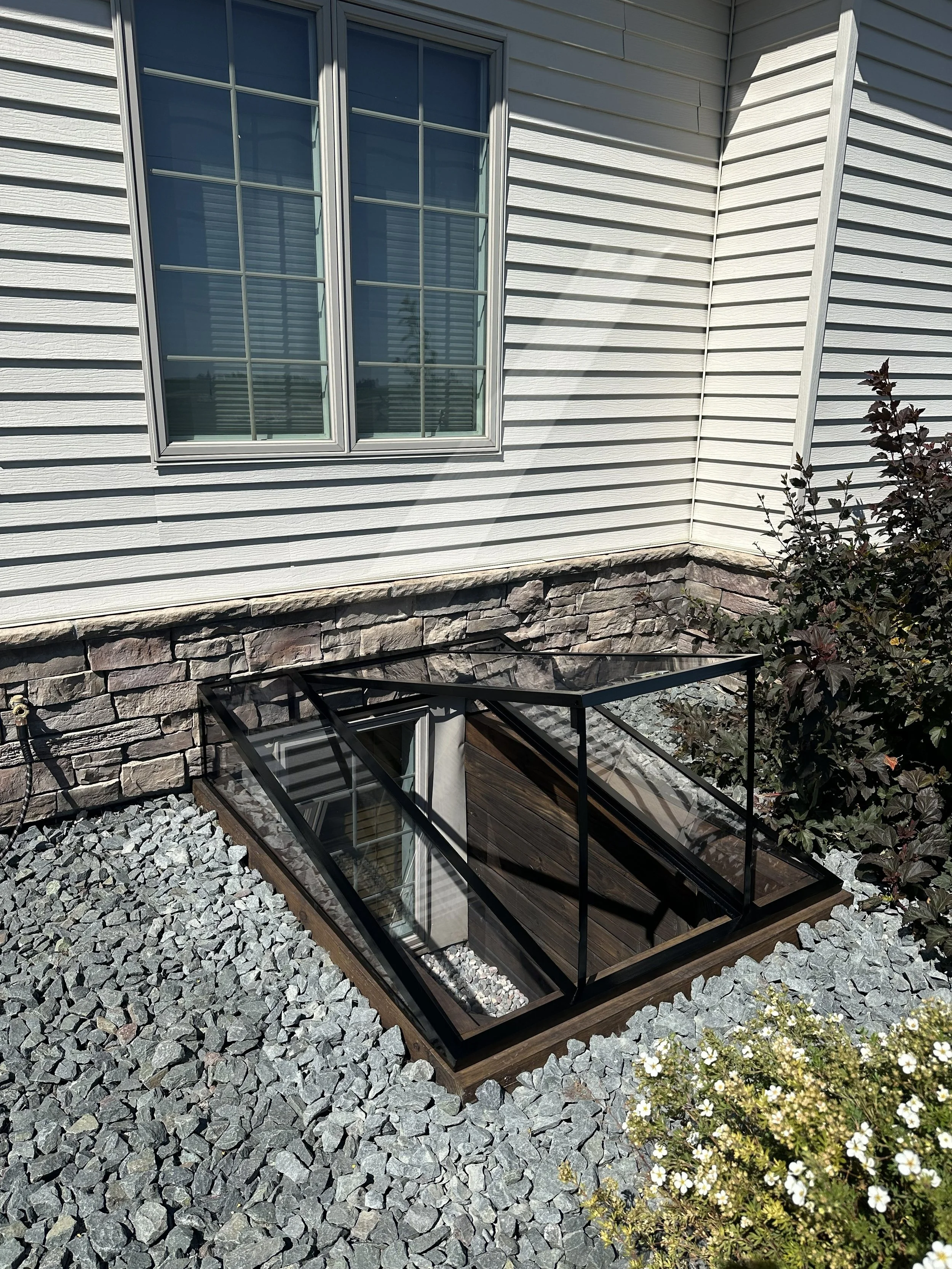 Exterior view of a house with white horizontal siding, a large window with multiple panes, and a stone foundation. There is a small glass-enclosed basement window and a landscaped area with gray stones and a flowering shrub.