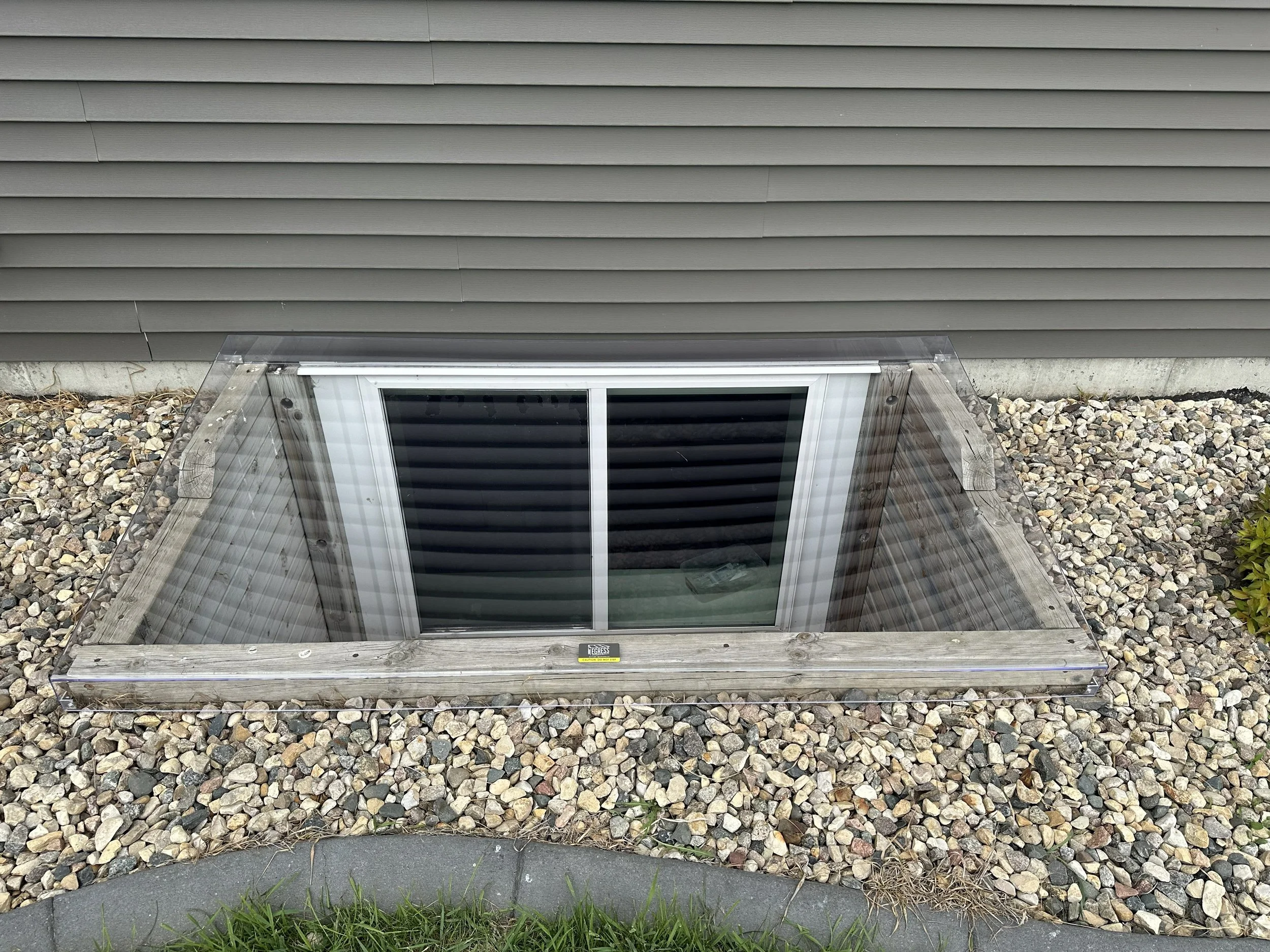 A basement window set into a house foundation, surrounded by a wooden frame, with a plastic cover and gravel outside.