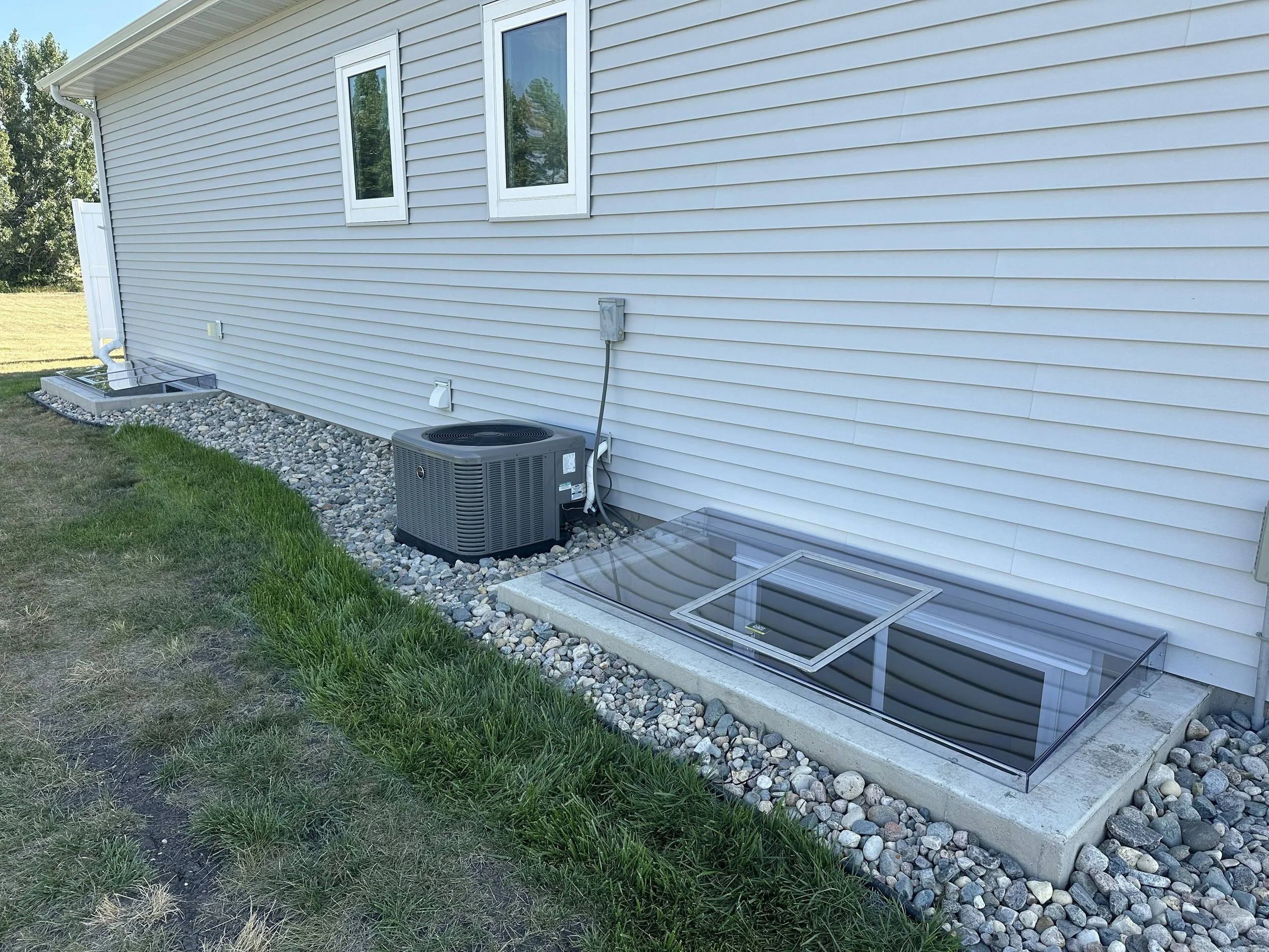 Back exterior of a house with white vinyl siding, two windows, an air conditioning unit, and basement windows with glass covers, surrounded by small rocks and a section of grass.