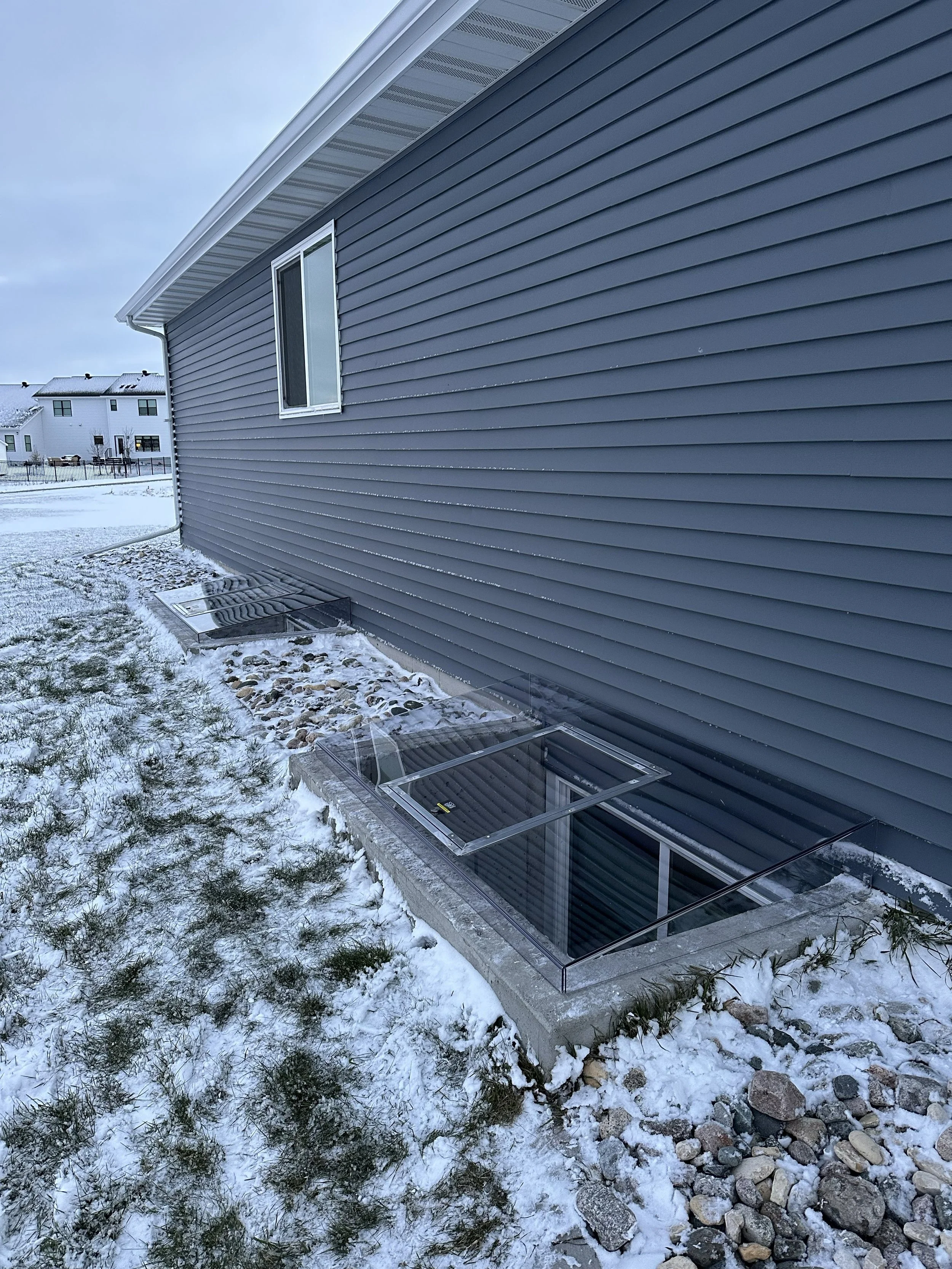 Side view of a house with gray siding and a window, snow-covered yard, rocks, and a small glass enclosure near the foundation, with neighboring houses in the background.