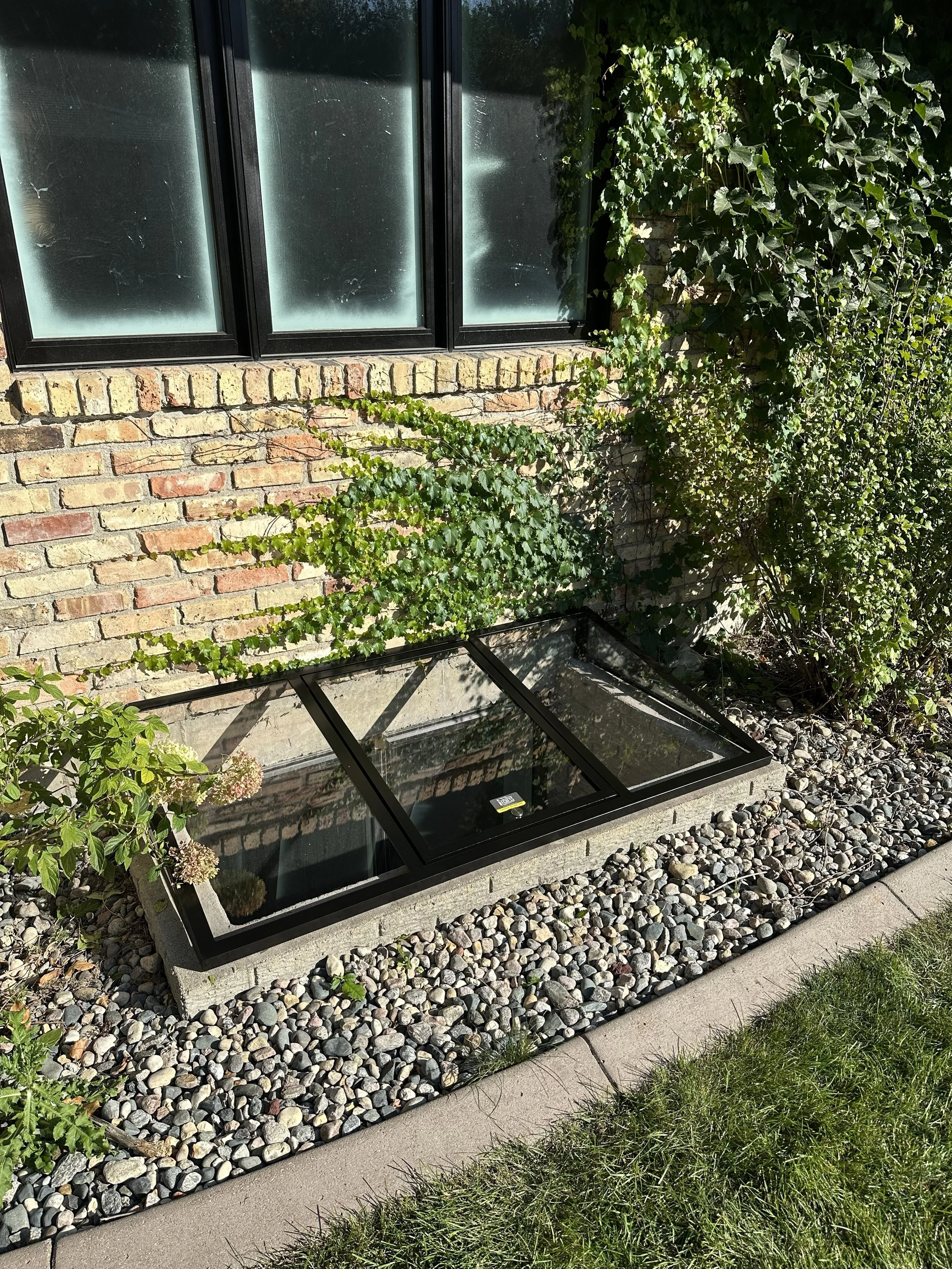 A brick house wall with first-floor window and surrounding plants. The window is partially opened, with one section lying flat on the ground. There are ivy and other green plants growing on the wall and beside the window, with a trampoline on the gra
