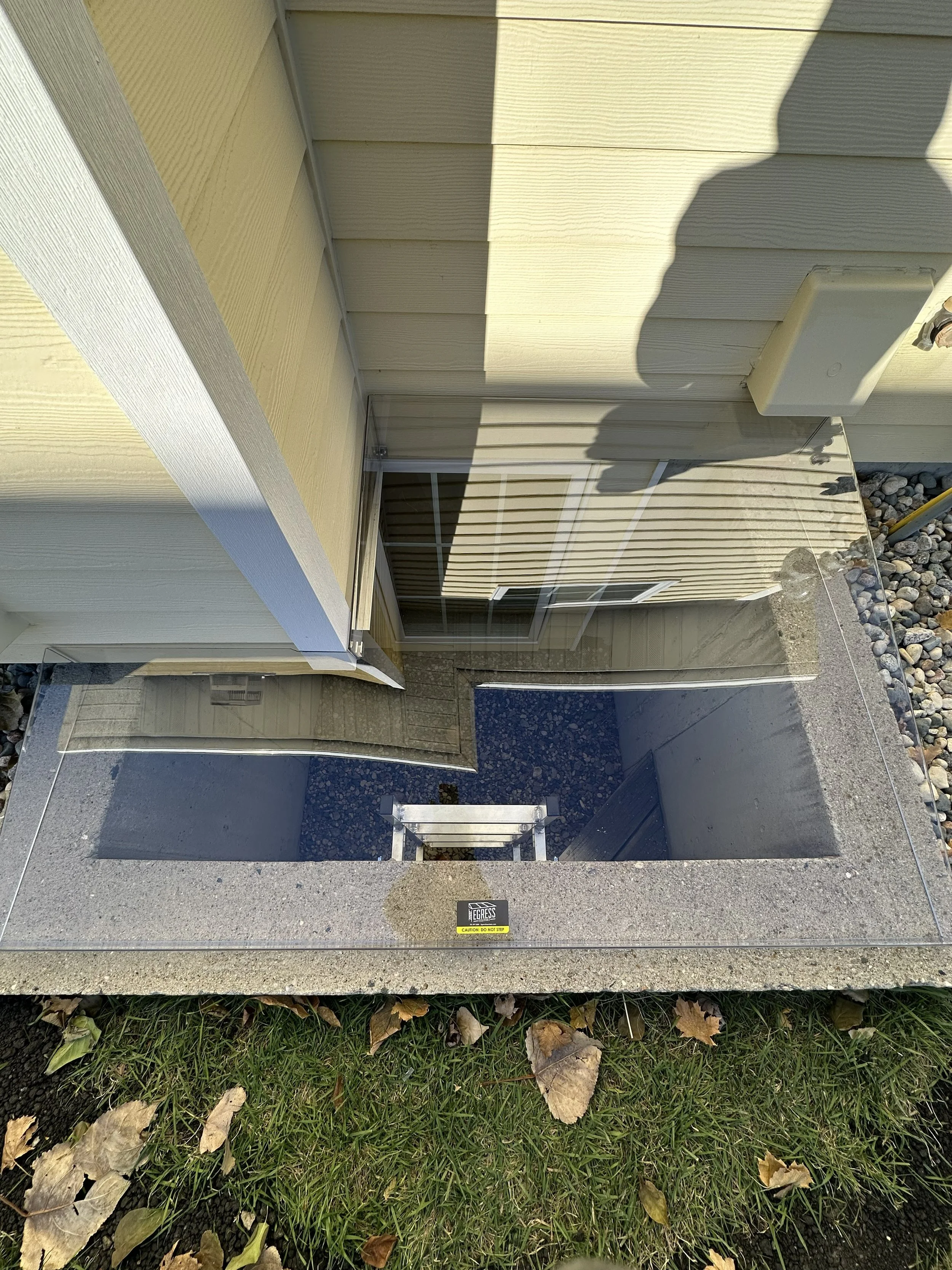 A concrete step leading to a building basement entrance with a safety warning sign and surrounded by fallen leaves and grass.