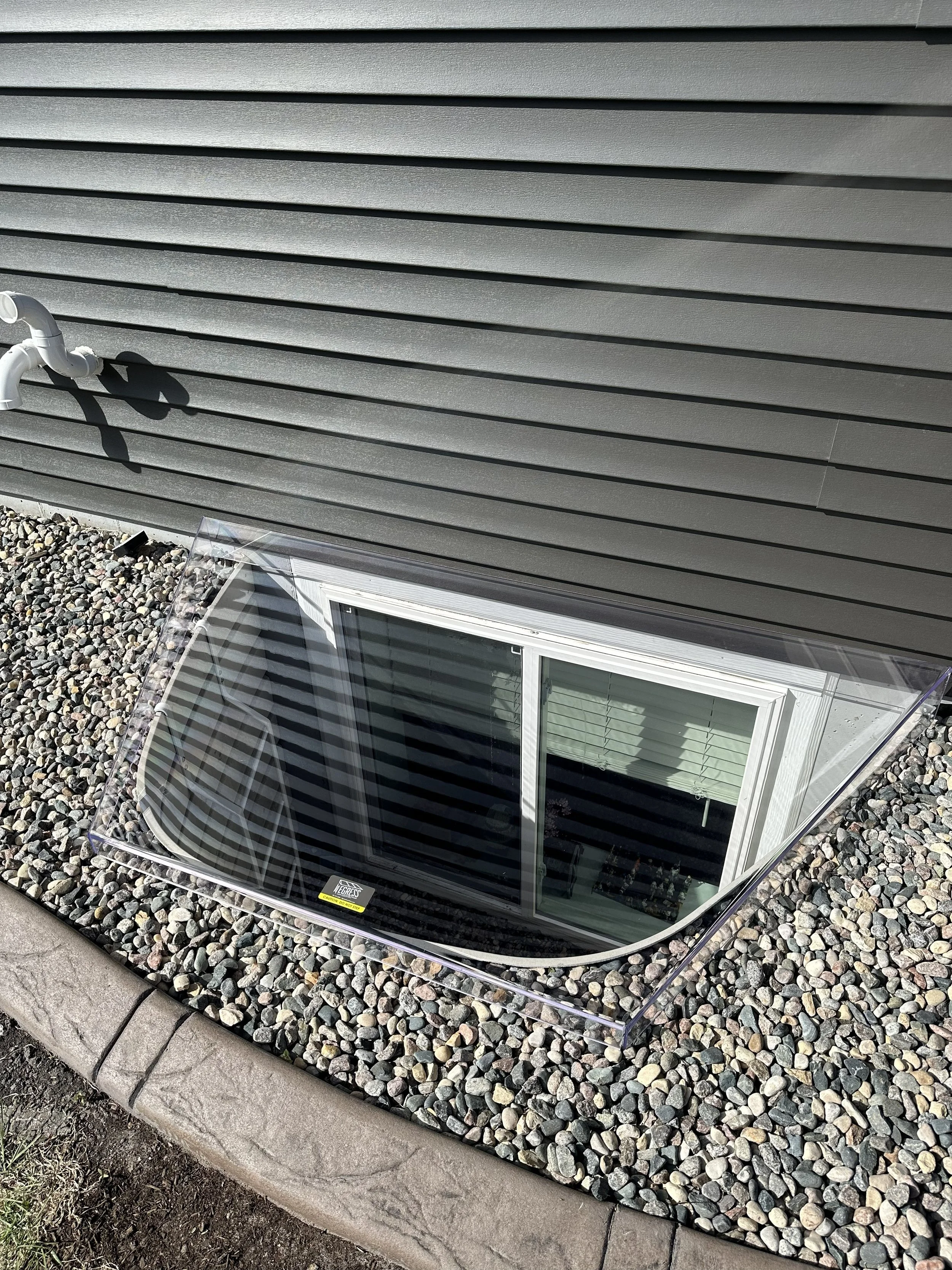 View of a brown house siding exterior wall with a white pipe and a small glass enclosed basement window on a gravel bed, with a concrete curb in the foreground.