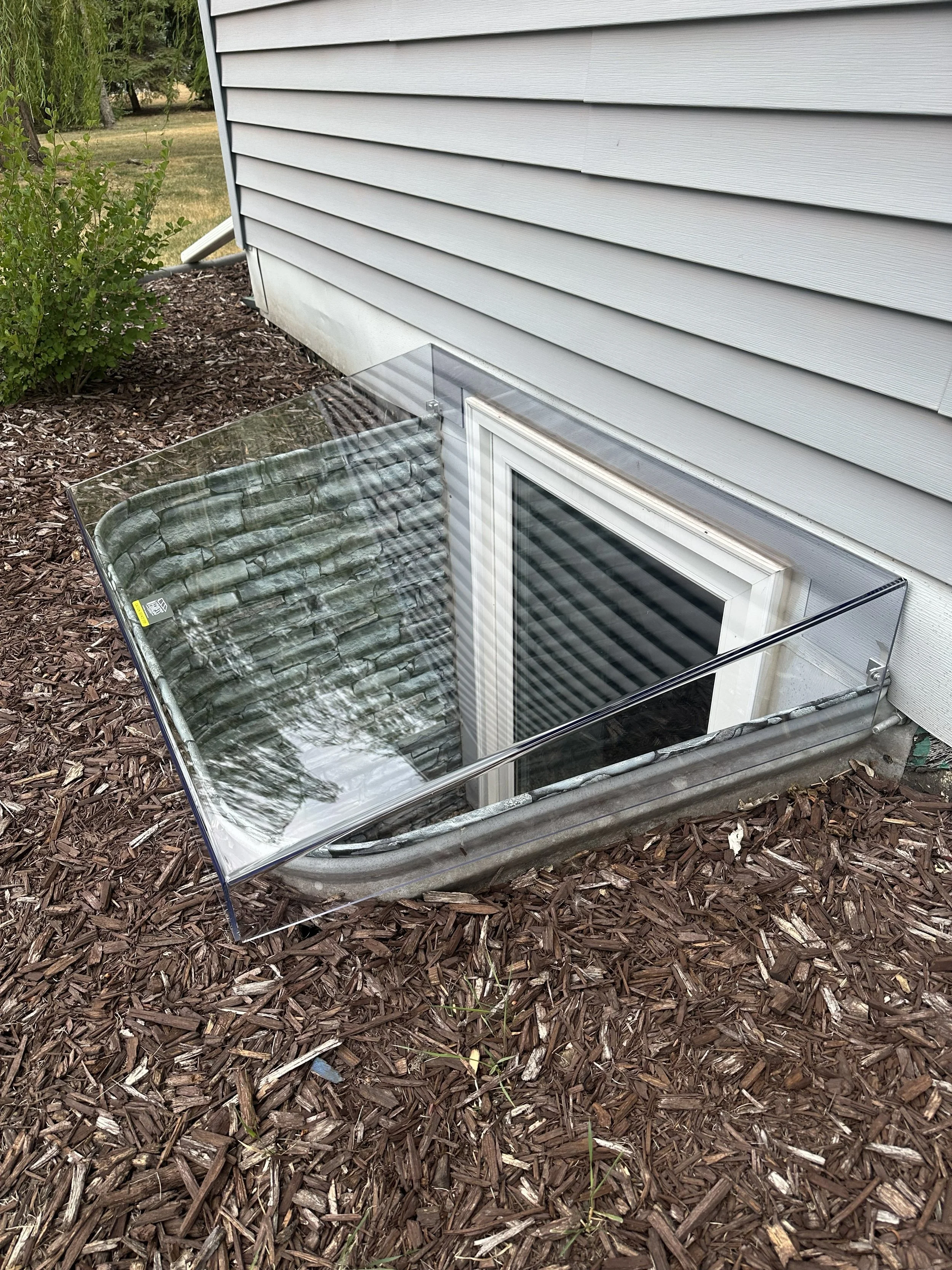 A basement window with a reflective glass cover installed in the ground outside of a house with grey siding, surrounded by mulch.