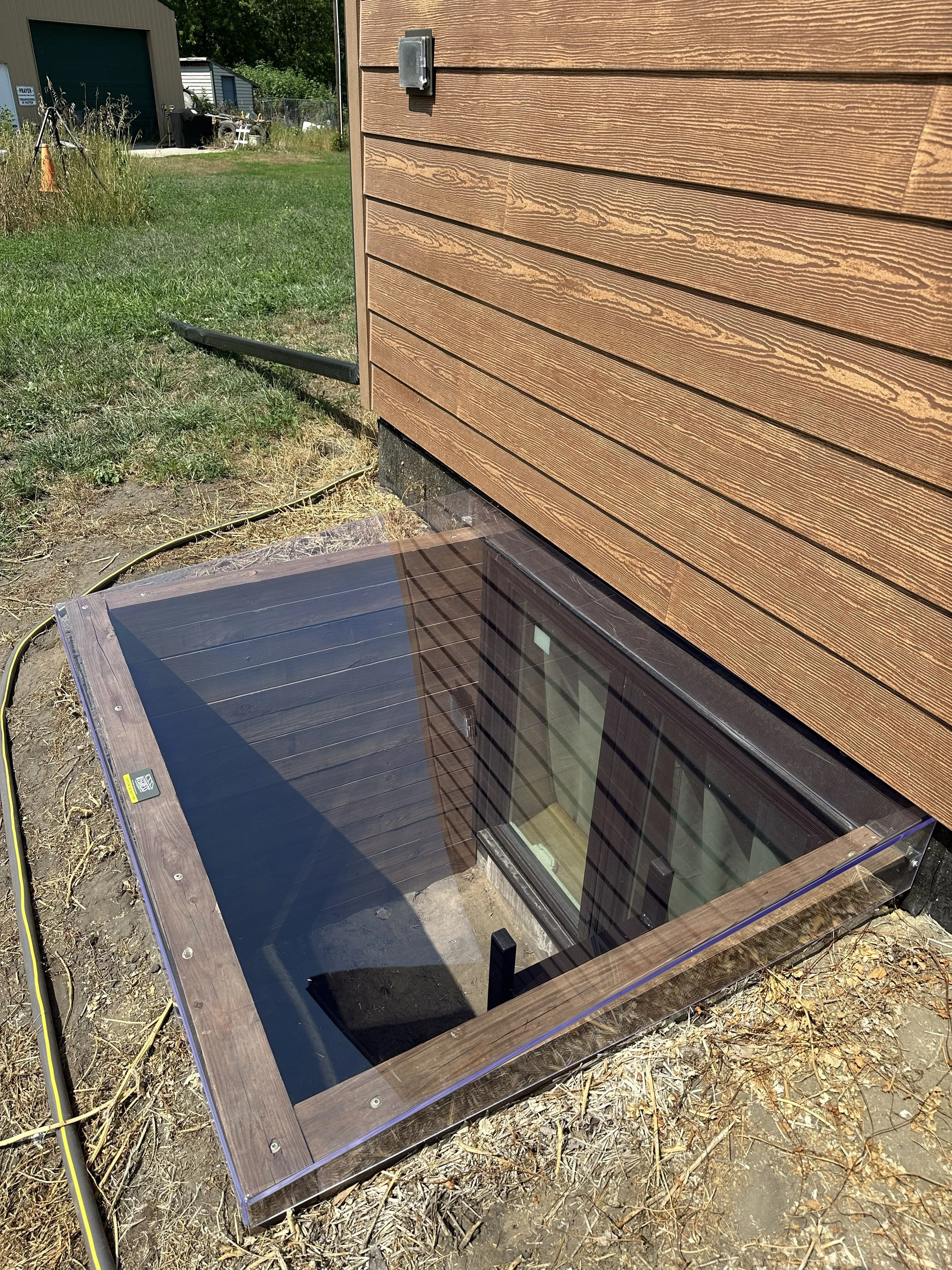 The image shows a corner of a house with brown horizontal wooden siding. A small, rectangular, gray electrical box is mounted on the wall. Below it, a window with a wooden frame and reflective glass is set into the wall. In front of the window, there