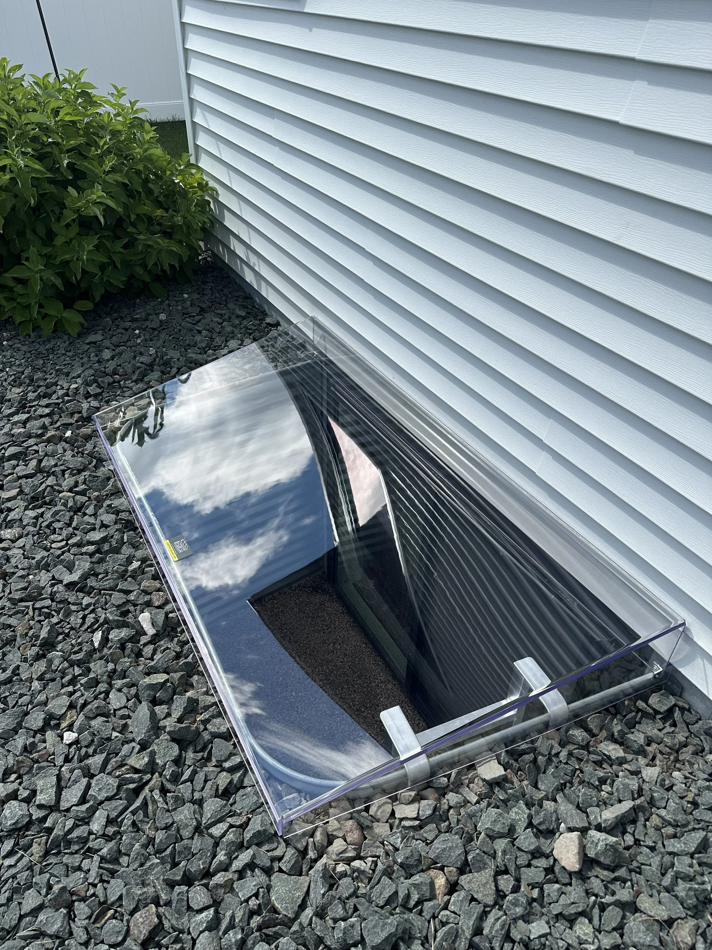 A reflective solar panel or mirror resting on gravel in front of a white house siding, with green bushes nearby.