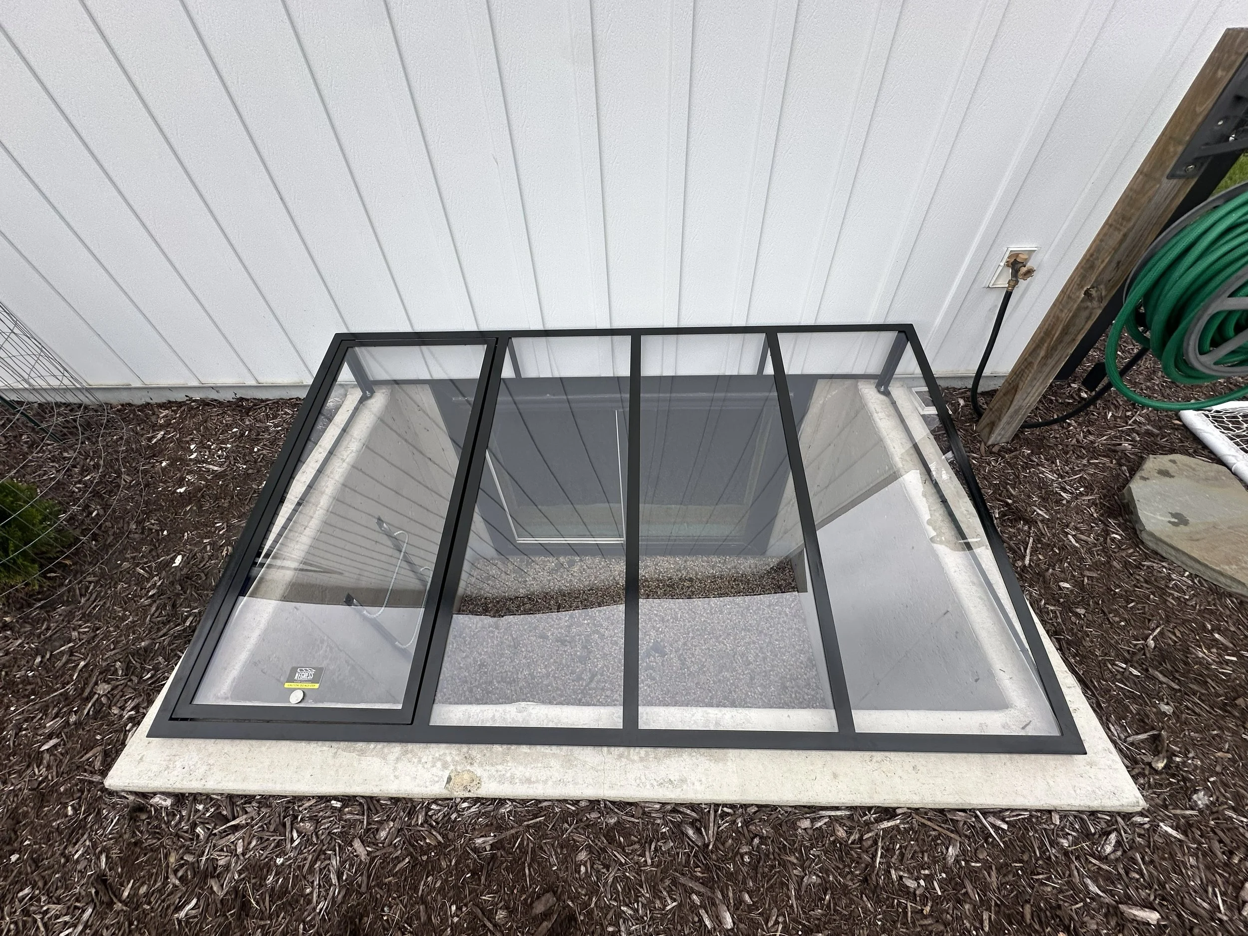 View of a small enclosed outdoor space with a glass door, outdoor garden hose, and soil mulch surrounding the area.