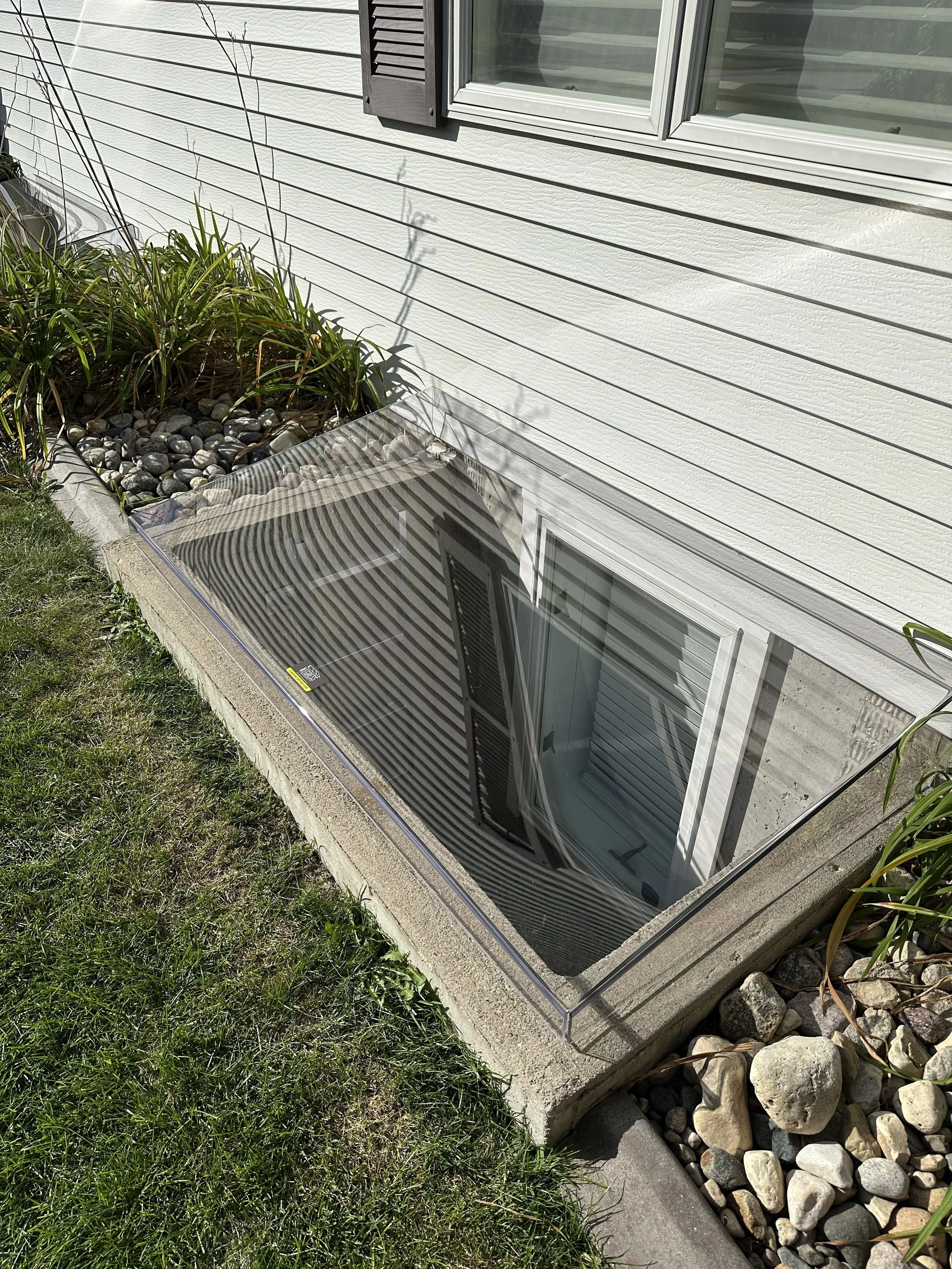 A basement window with a protective cover, surrounded by rocks, plants, and grass outside a house with white siding.