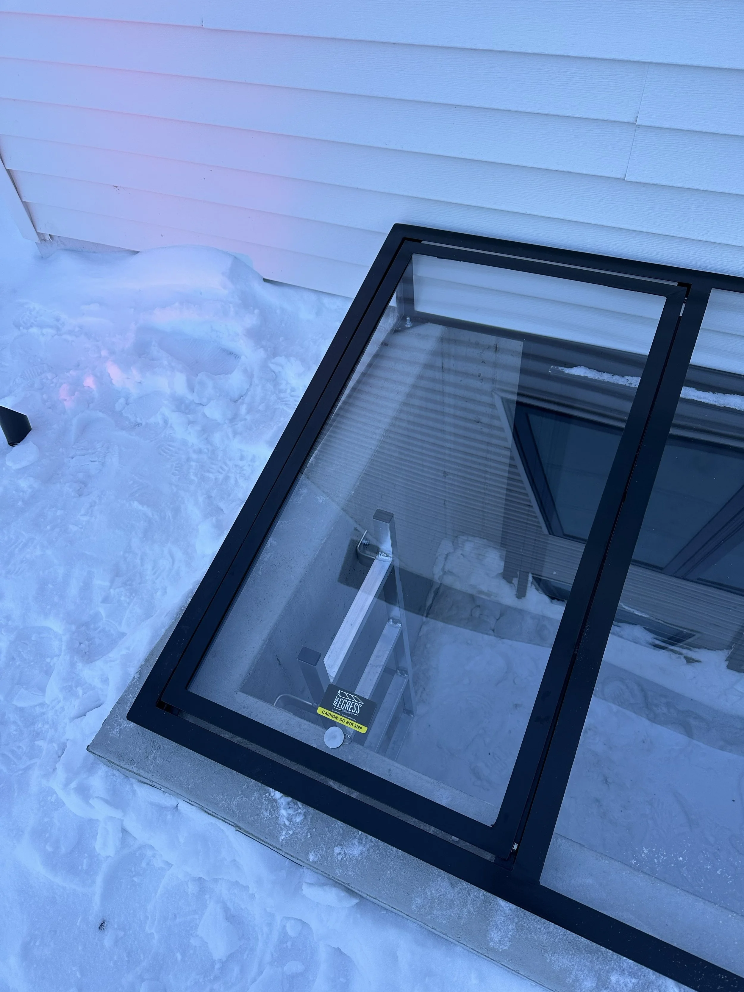 An outdoor scene with snow on the ground and a glass panel with a black frame and a metal stand inside. The glass panel has a yellow caution label and a small sticker reading 'Egress'.