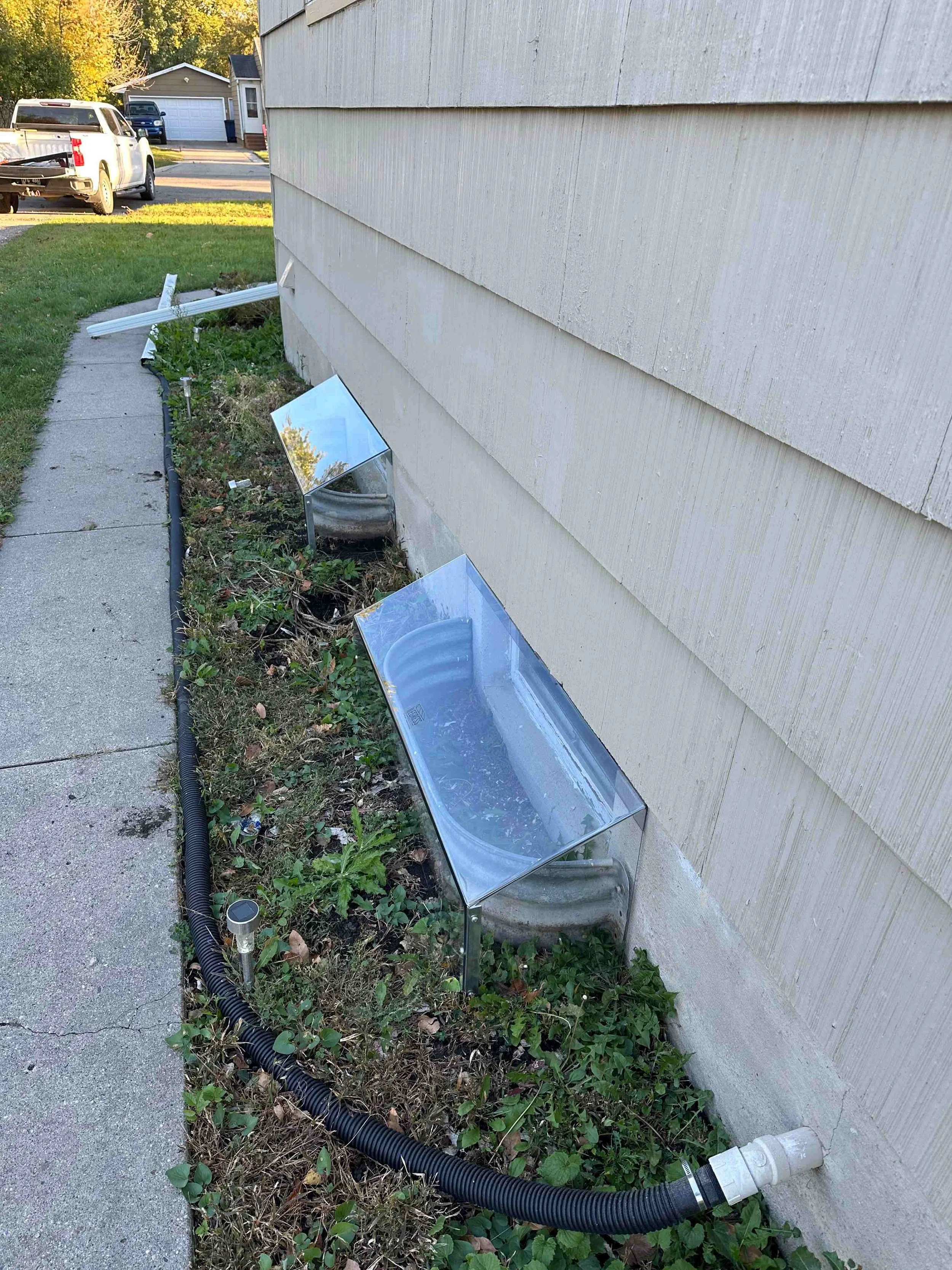 A house with beige siding. In the garden bed along the house, there are solar heat reflectors, a black corrugated pipe, some plants, and debris. The garden bed appears to have a bend in it, with a concrete walkway on the left side and a corner of a w