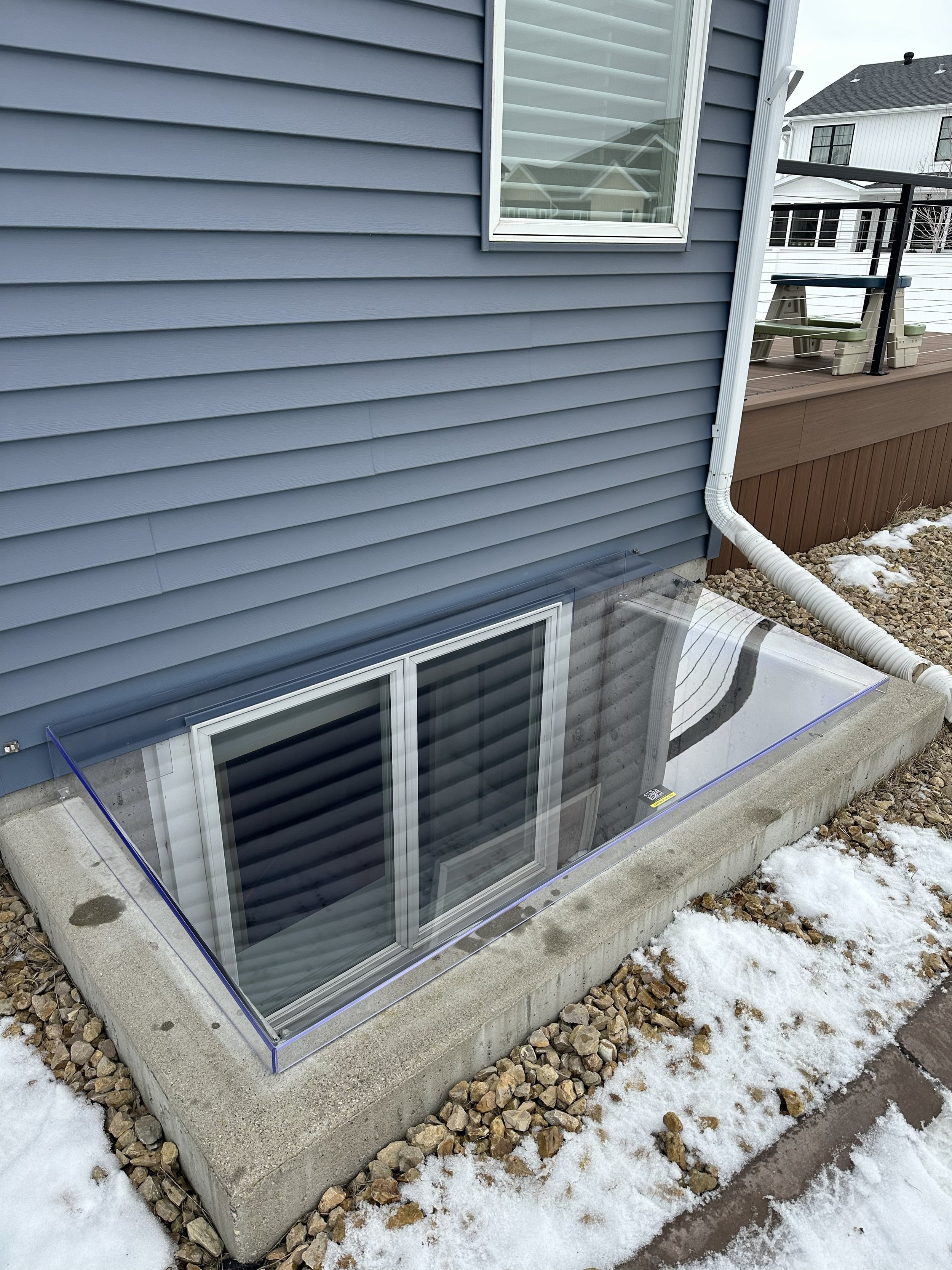 A basement window with a glass well installed outside a house with blue siding. The well is surrounded by gravel and some snow, with a concrete base and a railing on the deck above.