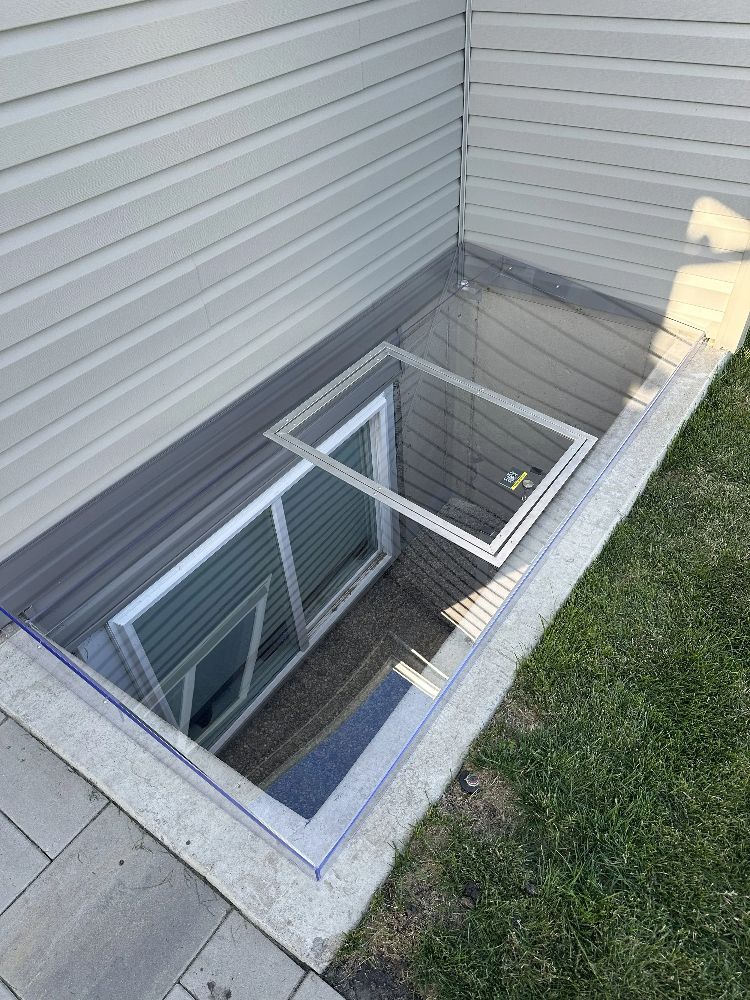 A glass basement window view of a house with beige siding and a concrete foundation, surrounded by grass and sidewalk.