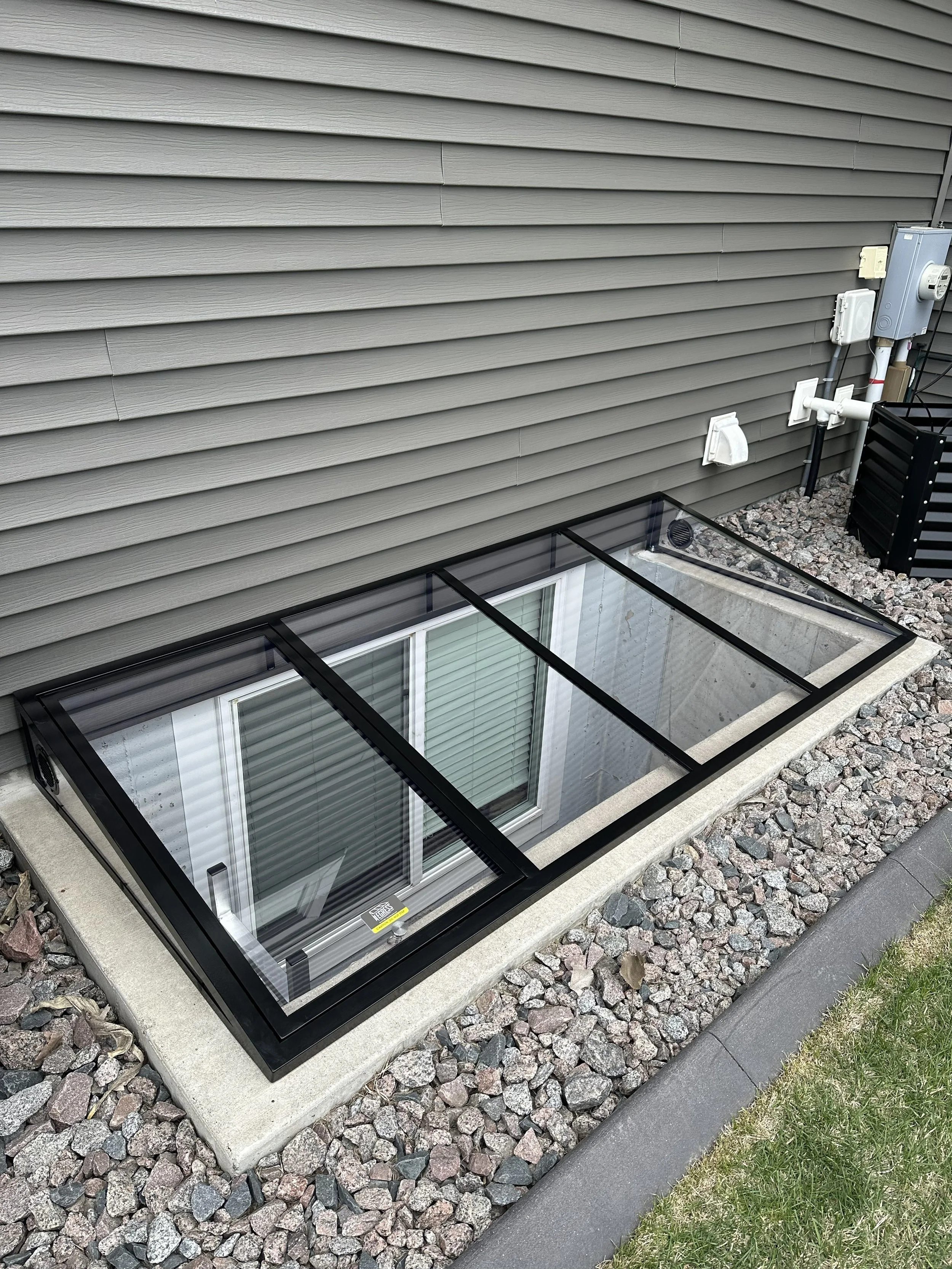 A basement window with a glass cover is set into a concrete foundation, surrounded by gravel, with exterior siding, a drain vent, and electrical boxes on the house wall, and sections of grass and pavement nearby.