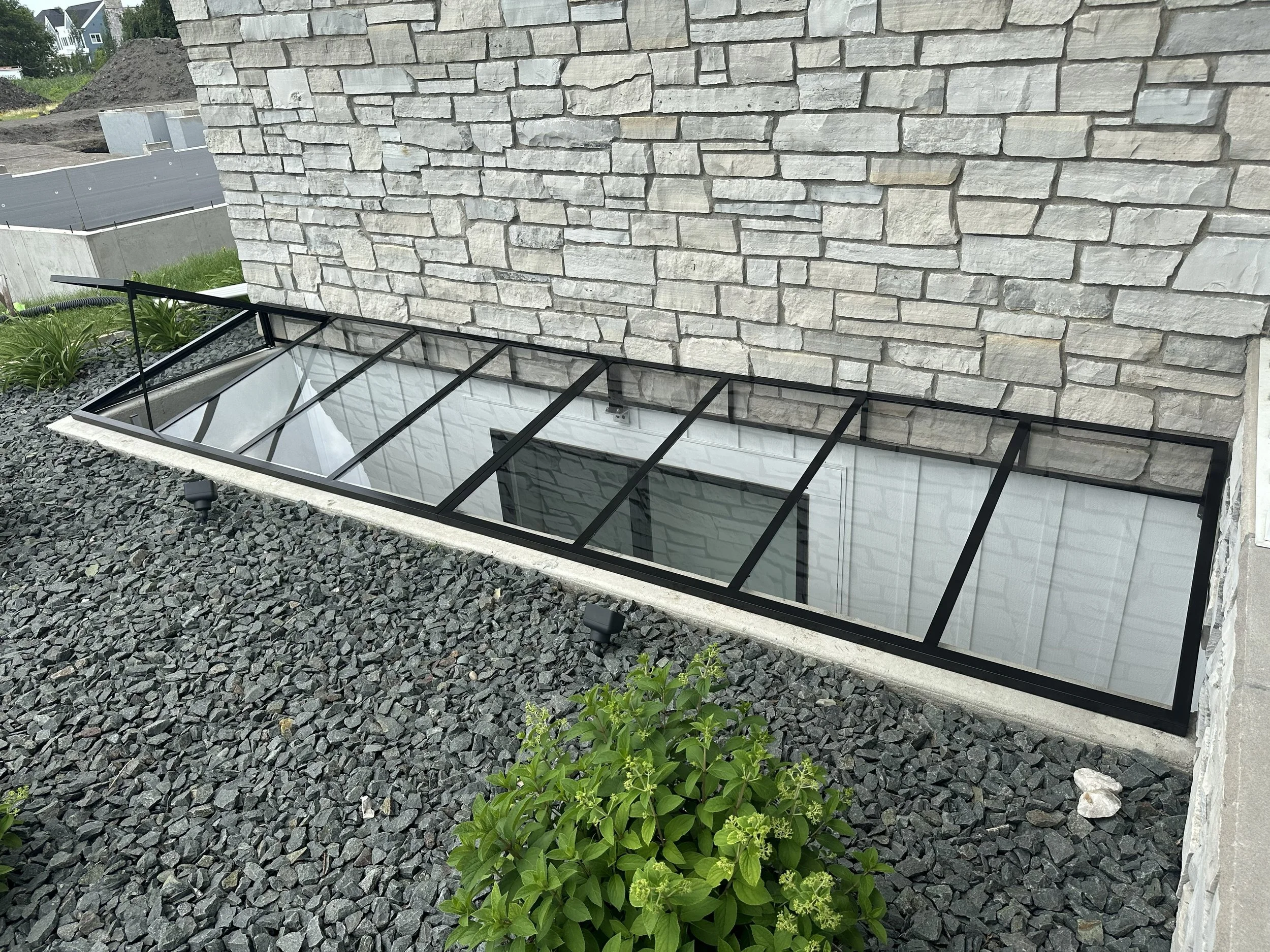 A rock garden with small black stones, a small green shrub, a large stone wall, a glass roof skylight, and a stone border.