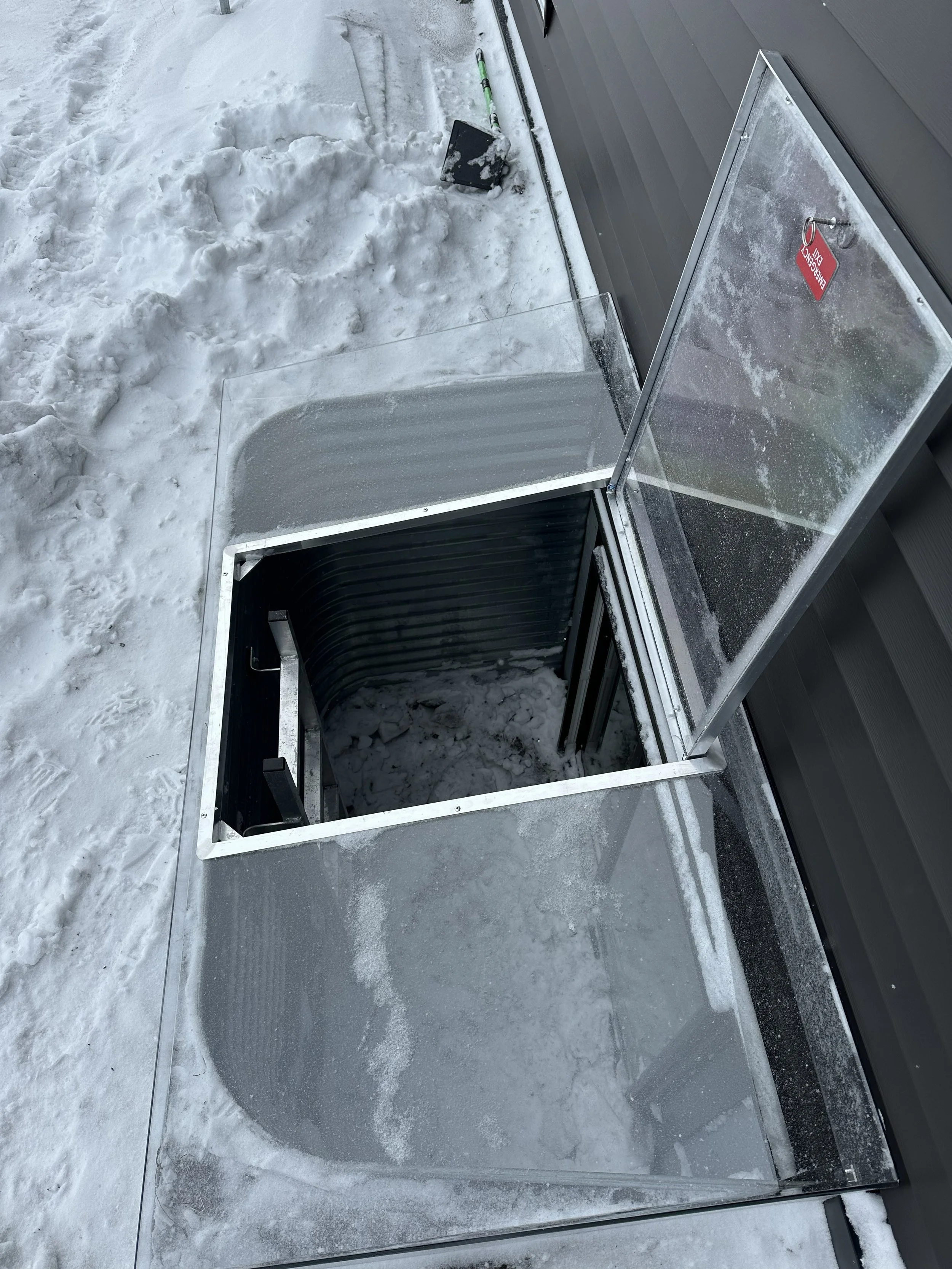 A snow-covered outdoor area shows a metal hatch with glass panels and a handle, partially open, revealing an interior space with a black lining. An ice scraper and snow shovel are also visible near the hatch.