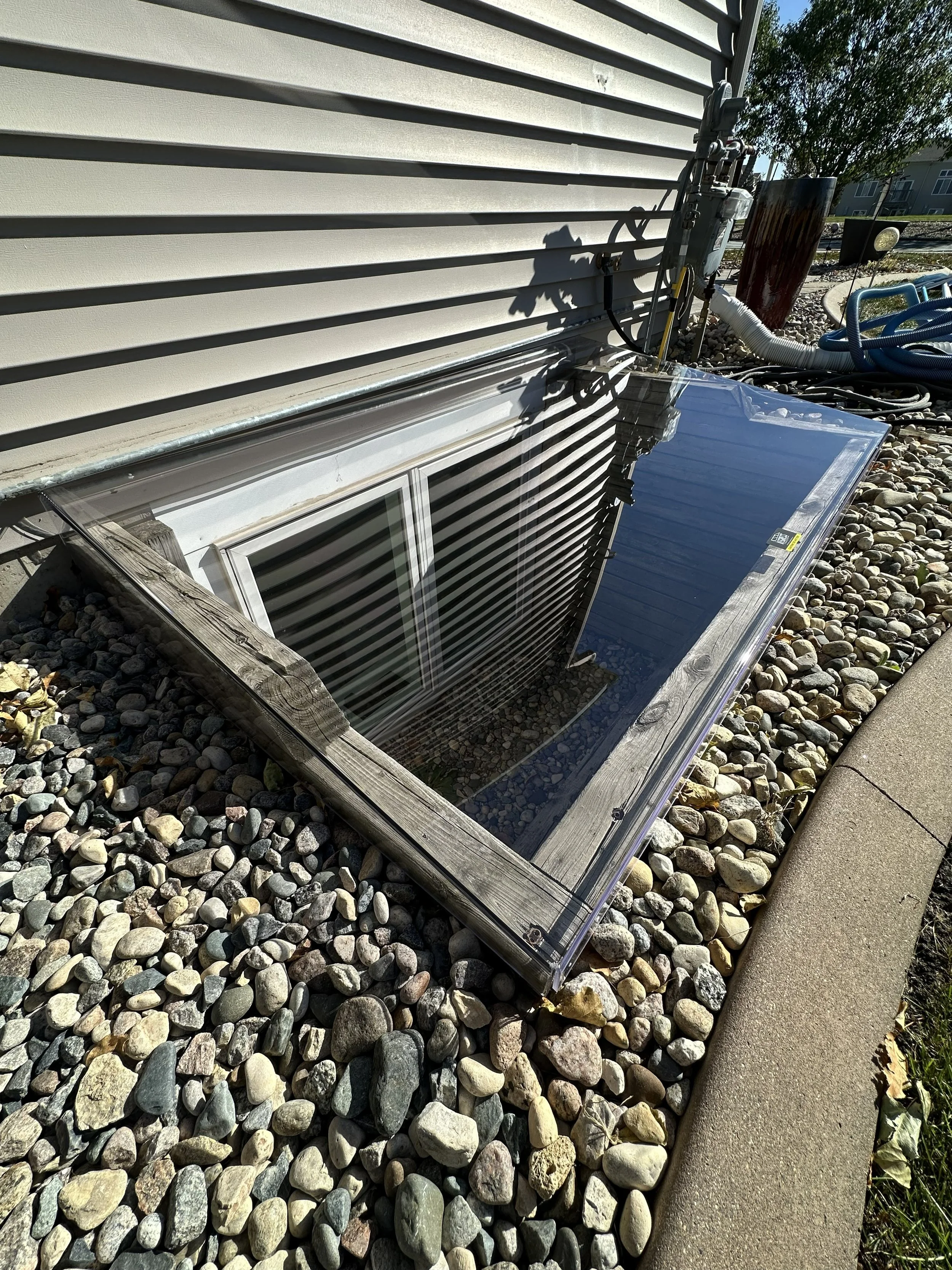 A basement window installed in a wooden frame, with a transparent plastic window well around it, surrounded by small multicolored rocks, next to a concrete sidewalk. There are pipes, electrical panels, and hoses nearby, and a tree with green leaves i