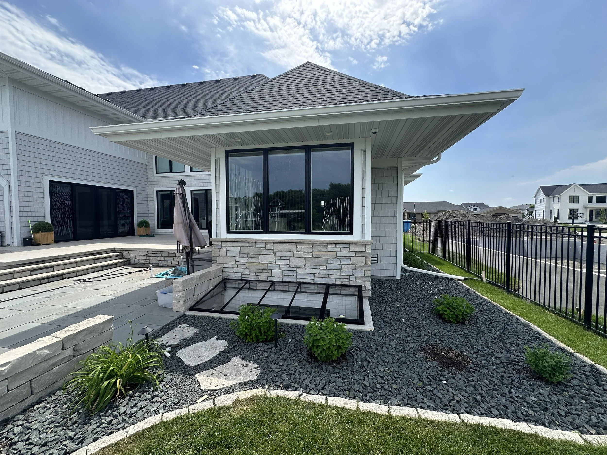 The image shows a modern house with a sloped shingled roof, large glass sliding doors, and a backyard patio with stone steps. The yard features landscaped elements including small plants, a bordered flower bed, black gravel, and a stone wall, with ne