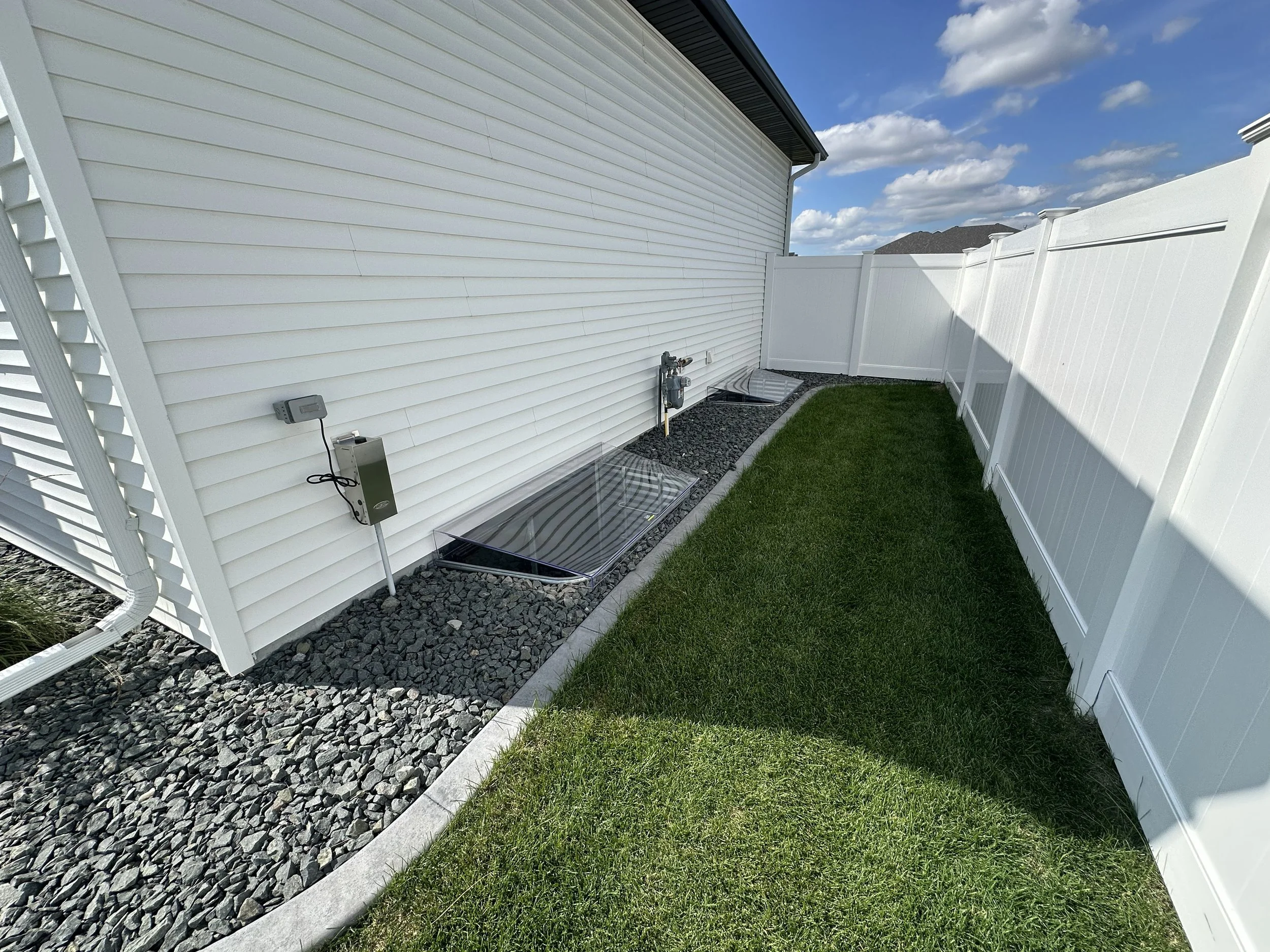 A backyard with white vinyl fencing, artificial grass, a gravel bed with rocks, and a white house with siding. The sky is blue with scattered clouds.