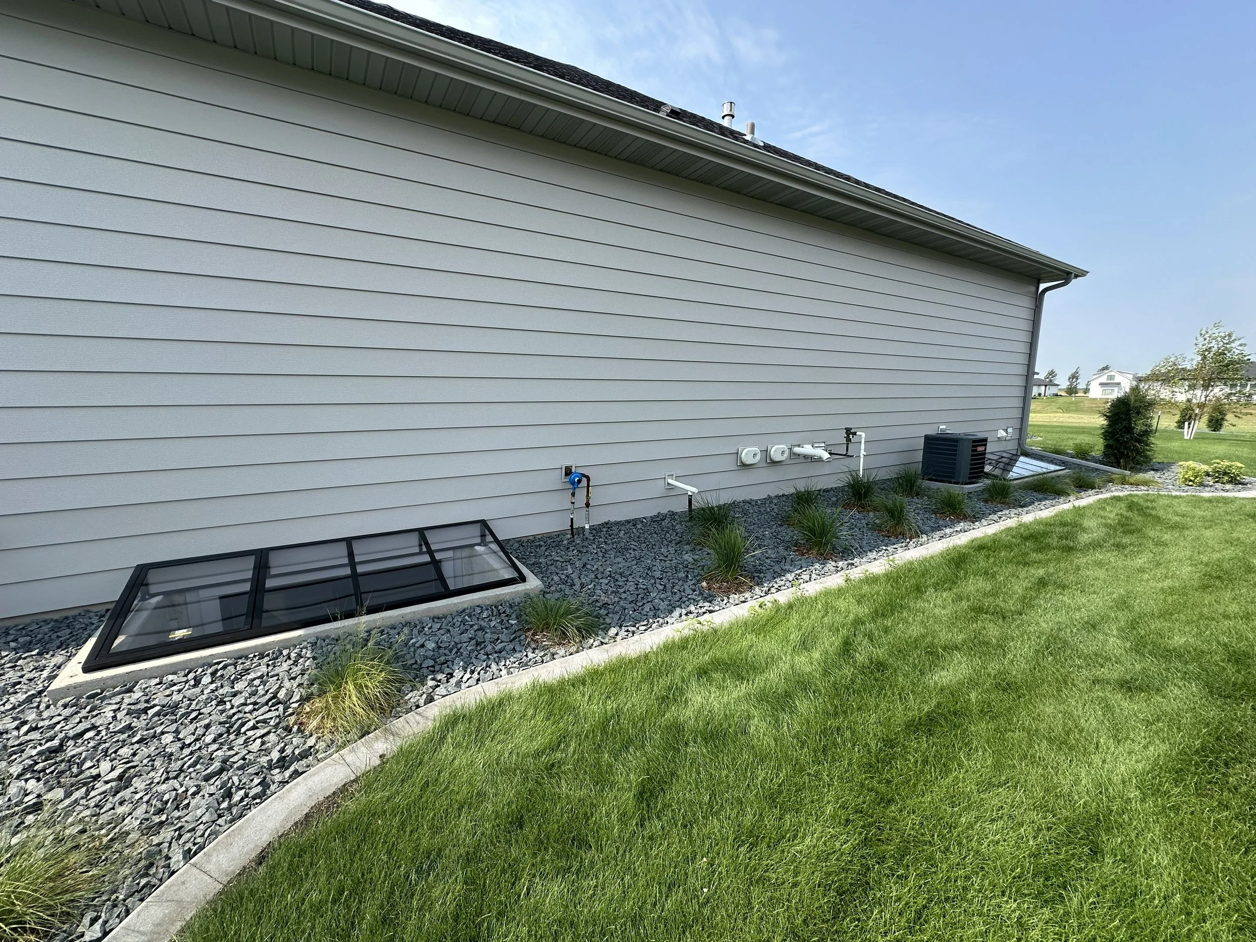 The side of a house showing beige horizontal siding, a vent, some piping, and a small landscaped area with rocks, grass, and plants, with neighboring houses and trees in the background under a blue sky.