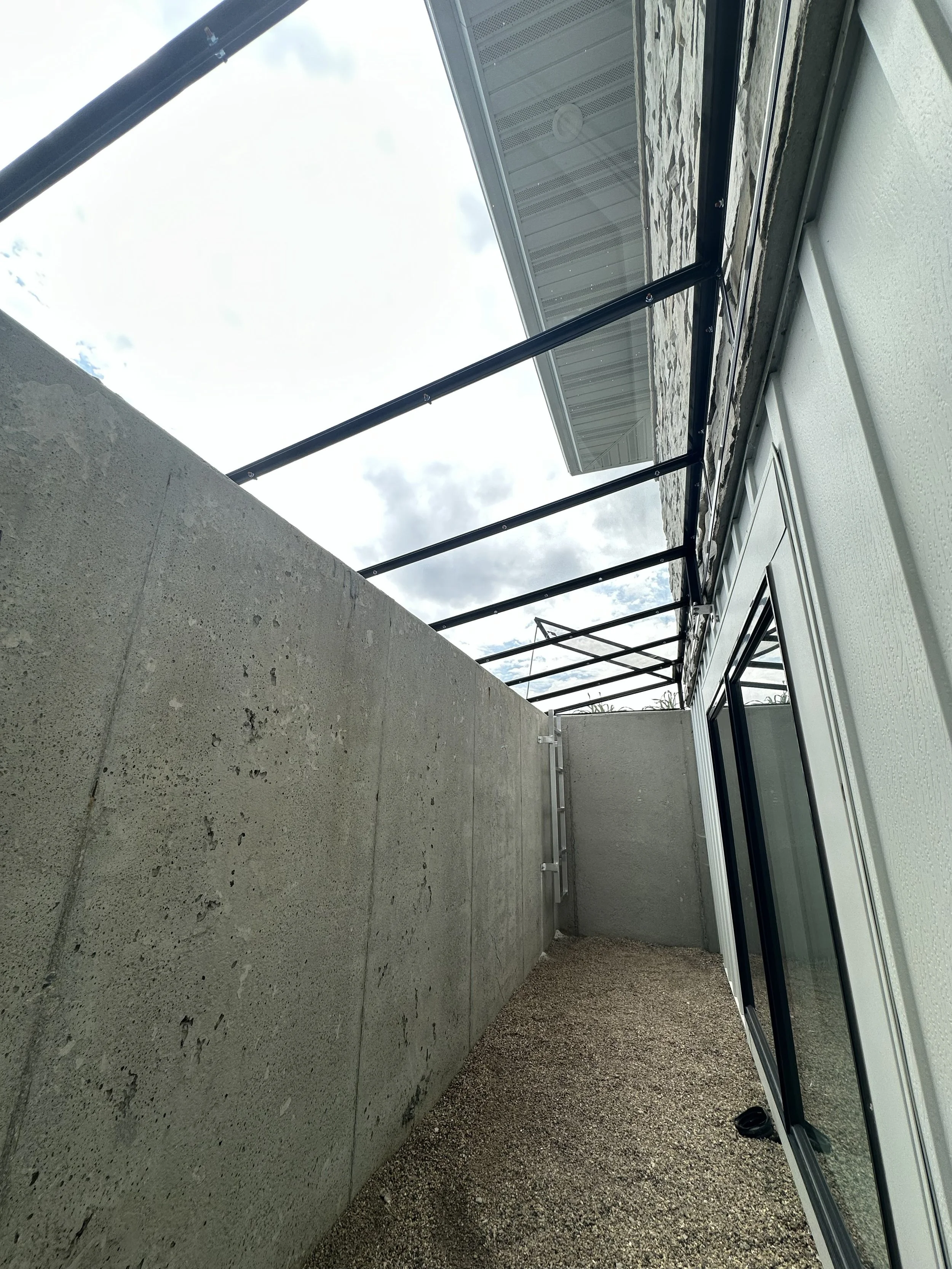 View of a balcony with a concrete wall on one side, gravel flooring, and a glass railing supported by black metal posts. Overcast sky with clouds is visible in the background.