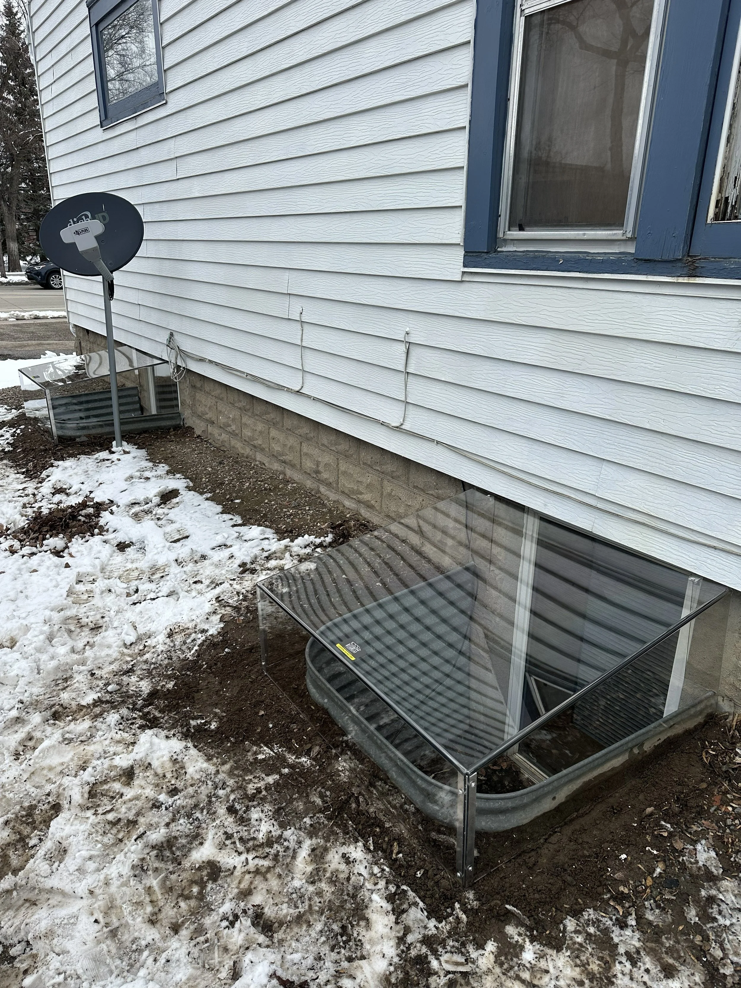 Backyard scene with snow-covered ground, a glass patio table, a satellite dish, and a house with white vinyl siding and a blue window frame.