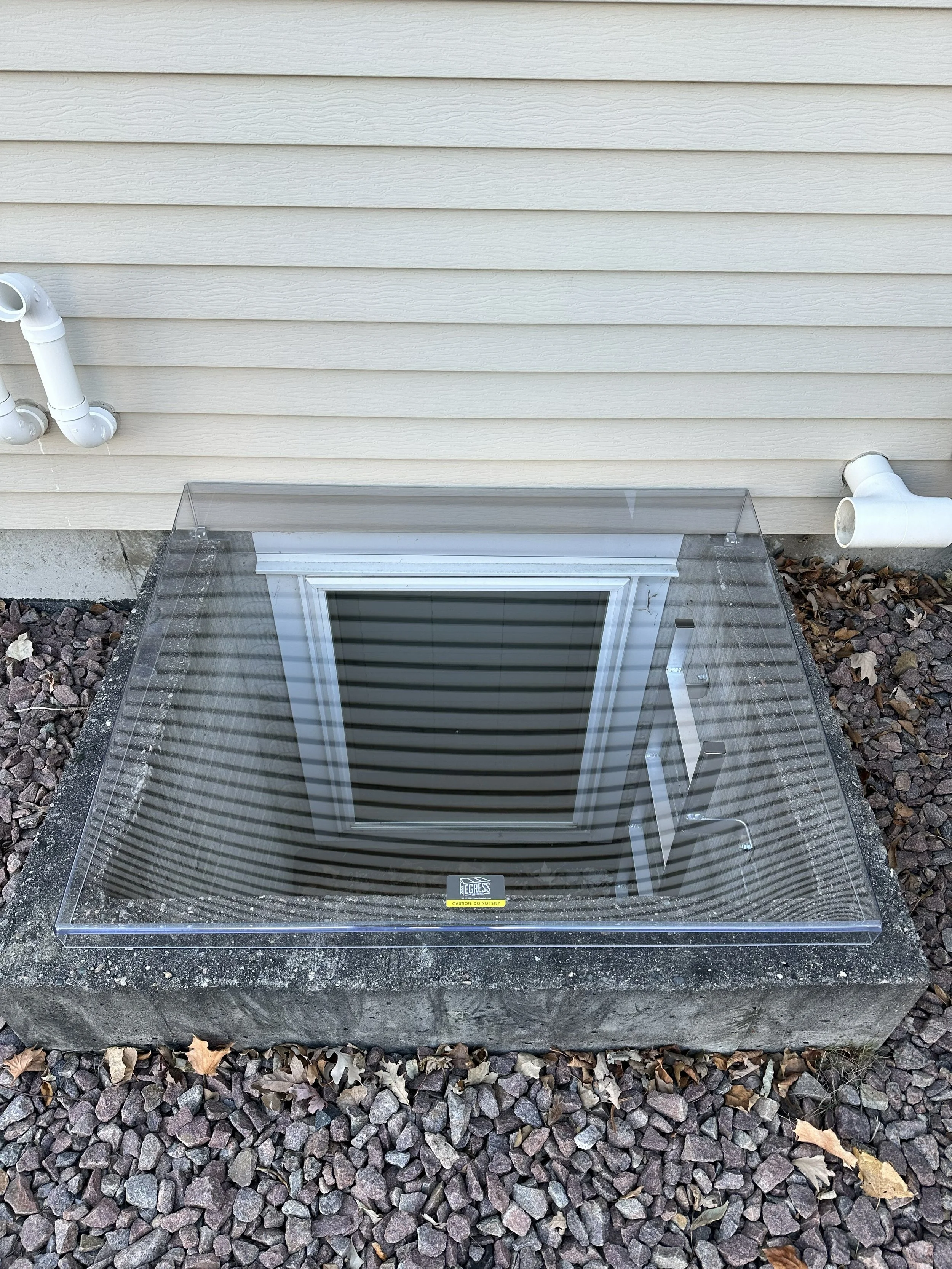 A basement window with a transparent plastic cover installed on a concrete foundation, surrounded by rocks and fallen leaves. The house has beige horizontal siding, and there are white drain pipes on either side of the window.
