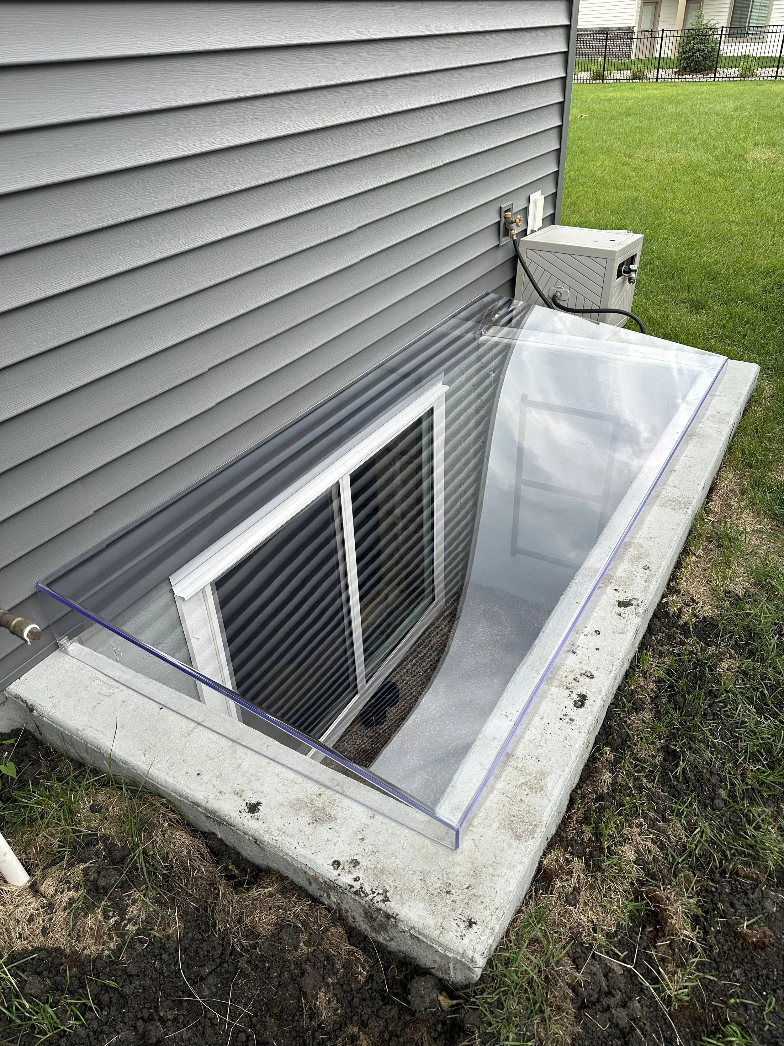 A house with gray vinyl siding and a small window below the ground level, surrounded by a concrete foundation. There is a glass window well cover and some grass and dirt in the yard.